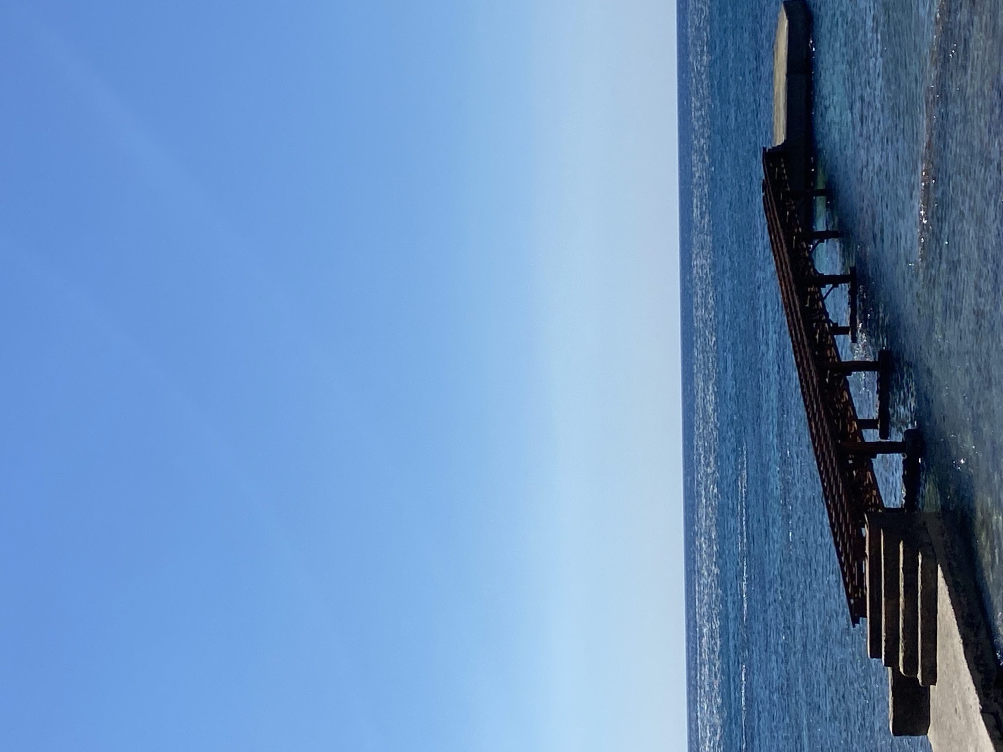 The image presents a serene coastal scene under bright daylight, located in Budva, Montenegro. The left two-thirds of the frame are dominated by a vast expanse of clear, cloudless blue sky, displaying a slight color gradient from brighter to a deeper blue. The right one-third features the deep blue sea, its surface gently rippling and reflecting bright sunlight.
Extending into the water from the bottom-right corner is a concrete or stone pier. Attached to this main structure and partially submerged in the clear, shallow water is a rusty metal structure, which appears to be a set of steps, a ladder, or a narrow ramp, supported by several vertical posts. Further along the pier, a prominent dark concrete block is visible, likely marking the end of a breakwater or a section of the pier itself. The water near the structures is clear enough to reveal glimpses of the seabed.
The scene is devoid of human activity, conveying a sense of peace and stillness. The bright, direct illumination suggests it is mid-morning or early afternoon on a sunny day with clear weather. There is no visible text in the image.