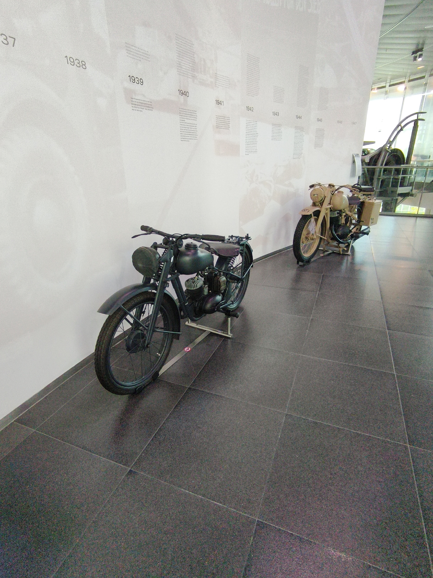 The image shows a display of vintage motorcycles within a museum setting. On the left, a dark-colored motorcycle is parked on a dark tiled floor, with its front wheel and fender prominently visible. To the right, a lighter-colored motorcycle with a luggage rack is also on display, supported by stands. The background features a large, curved wall with a timeline of years and text, suggesting an exhibition on historical events. The lighting is bright, illuminating the motorcycles and the exhibition wall. The overall environment appears to be a spacious, modern museum hall with polished dark flooring and a stark white curved wall that serves as a backdrop for historical information. The image is taken from a low angle, emphasizing the motorcycles.