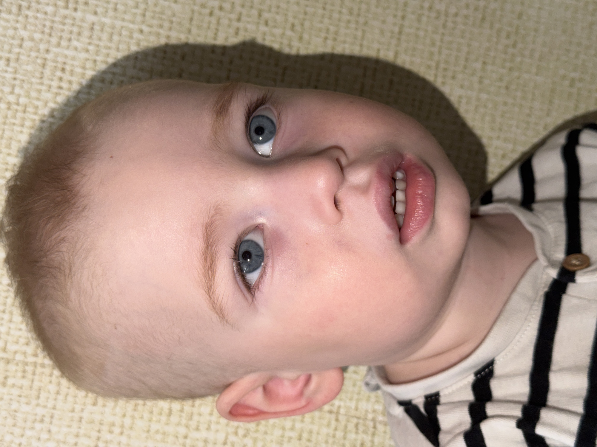 A close-up shot depicts a baby lying on a textured surface, possibly a blanket or sheet. The baby has fair skin, light blonde hair, and striking blue eyes. The baby's mouth is slightly open, revealing some teeth. The baby is wearing a striped shirt with a button visible near the neck. A shadow falls across the upper left portion of the baby's head, suggesting a light source from the side. There is no evidence to suggest the location is Chaikovsky, Russia.