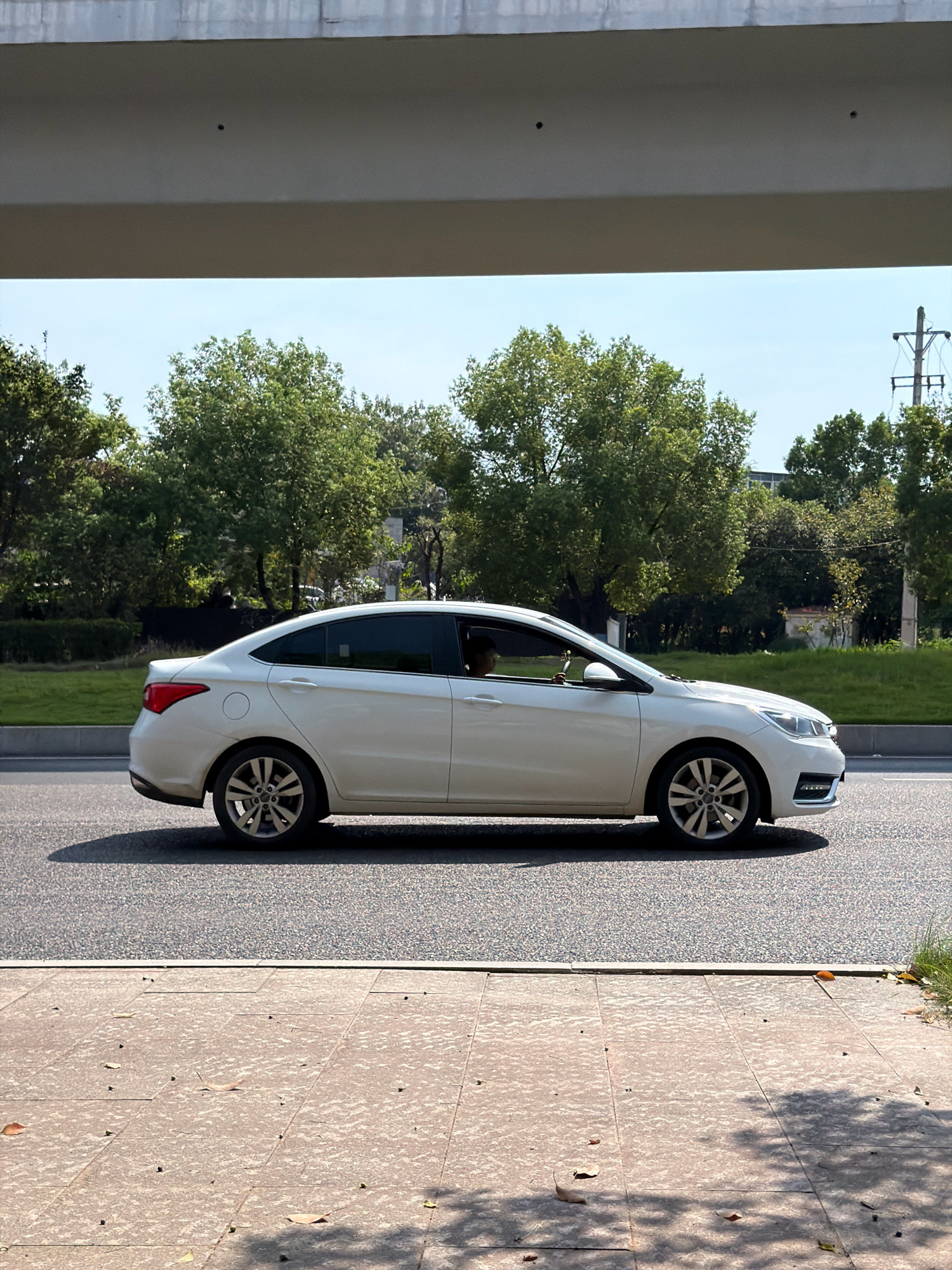A white sedan is driving on a paved road in Jiangjin, China. The car is seen in profile, moving from left to right. The driver, partially visible, is looking forward. Lush green trees line the background, and a concrete overpass is visible overhead, casting shadows on the road and sidewalk. The sky is clear and blue. The sidewalk in the foreground is made of light-colored square tiles with fallen leaves scattered on it. A shadow of tree branches falls across the sidewalk.