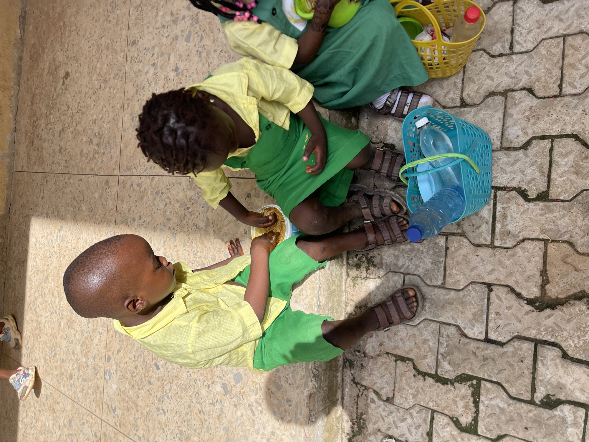 An overhead shot captures three young children sharing a meal outdoors in Akwanga, Nigeria, under bright daylight. In the foreground, a young boy with a bald head, wearing a yellow checkered shirt and green shorts, sits on a light-colored speckled tiled floor, looking towards a bowl of food. His right hand is near the bowl, suggesting he is reaching for or being offered the food.
Centrally, a young girl with dark, braided hair and small beads sits opposite the boy. She is also dressed in a yellow checkered shirt and a green pinafore dress. She holds a white bowl filled with light-colored grains, possibly corn or a similar staple, which she appears to be sharing with the boy. Her bare feet, clad in brown sandals marked "ZEBRA," rest on a patch of grey interlocking pavers.
Partially visible in the background, another young girl in a green dress is seated behind the central girl, also on the tiled surface. A yellow plastic basket containing a red-capped bottle and some white items is placed near her. Adjacent to the central girl's feet, a blue plastic basket holds two clear water bottles. The scene combines two distinct ground textures: the smooth, speckled tiles and the rougher, moss-flecked interlocking pavers, illuminated by direct sunlight creating distinct shadows. The children's attire suggests school uniforms, implying the activity might be a snack or lunch break.