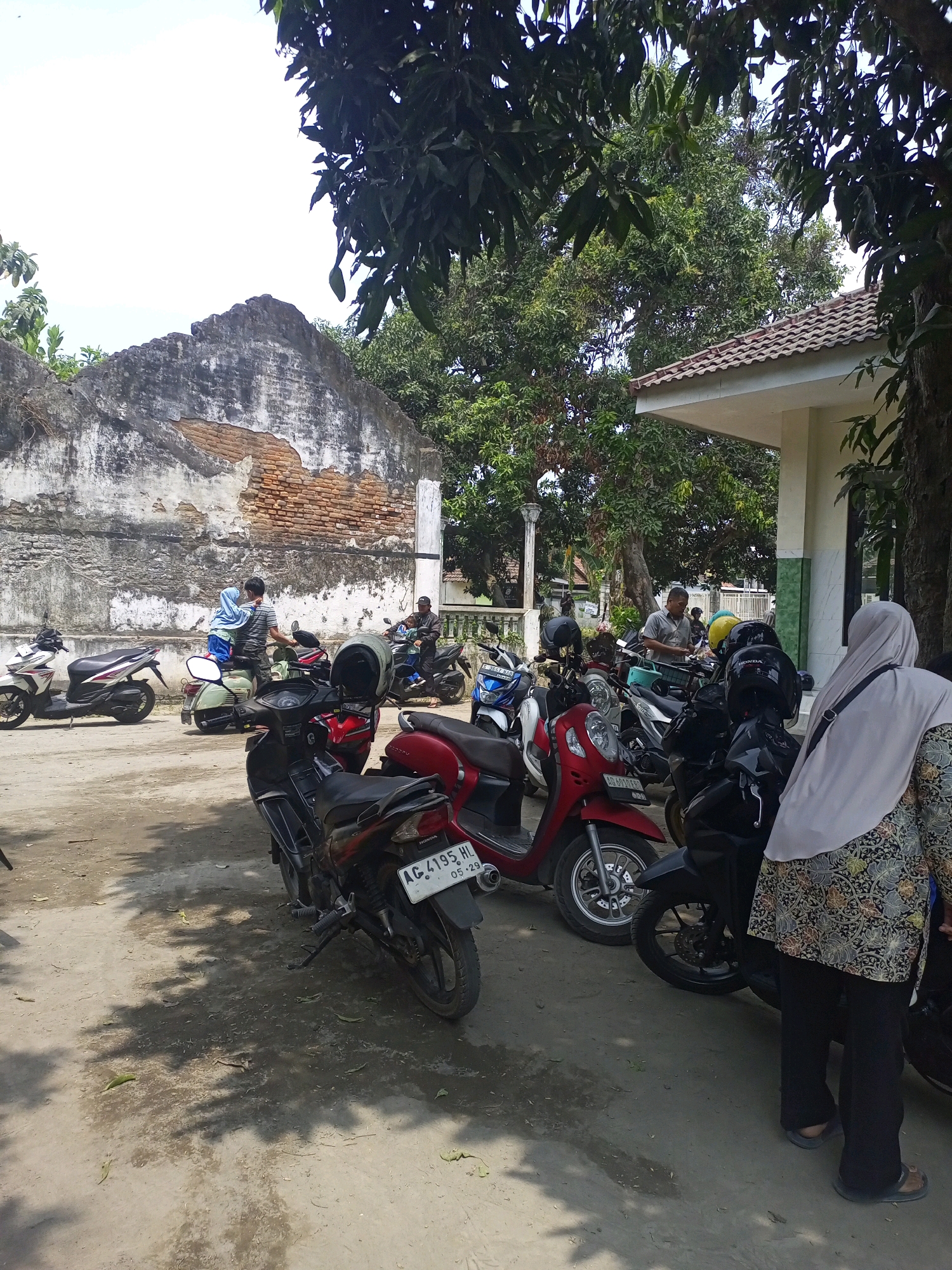 In the city of Gurah, Indonesia, a collection of parked motorcycles is visible in an outdoor area with a dirt and gravel ground surface. The scene is set against the backdrop of a partially ruined brick building with weathered walls on the left and a modern building with a tiled roof on the right.
Several individuals are present. In the foreground, a person wearing a light-colored hijab and a patterned top is standing next to a motorcycle. To their left, a dark-colored motorcycle with a license plate reading "AG 4195 HL 05-29" is parked. Further left, a red scooter is parked, followed by a dark motorcycle.
In the mid-ground, a man in a striped shirt is standing next to a light green scooter, with a child wearing a blue headscarf seated on the scooter's rear. Behind them, other motorcycles are parked, including a white and red scooter on the far left. Two men are also visible in the mid-ground, one standing and one seated on a motorcycle.
The environment is characterized by trees and foliage, with dappled sunlight creating shadows on the ground. The weather appears to be clear and sunny.