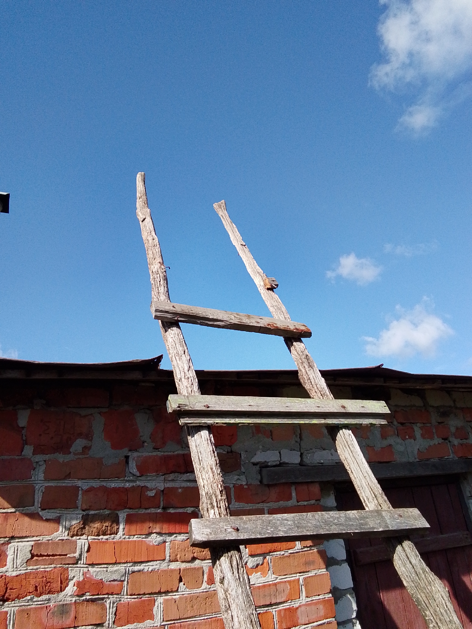 The image displays a rustic, handmade wooden ladder leaning against a weathered brick wall under a bright blue sky, characteristic of Khlevnoye, Russia.
The primary subject is a tall, roughly constructed ladder. Its two vertical supports are irregular, unrefined logs or branches, showing the texture of natural wood and wear. Several horizontal planks serve as rungs, affixed to the main supports with visible nails. The wood of the ladder appears aged and bleached by the sun, suggesting long-term outdoor exposure.
Behind the ladder stands a substantial wall made of red bricks. The mortar between the bricks is highly visible, exhibiting a mix of grey, white, and reddish hues, indicating various stages of construction or repair. The brickwork itself shows signs of age and weathering. Above the brick wall, a dark, curved roof edge, possibly made of corrugated material, is partially visible. To the lower right, a section of a dark wooden door or shutter is visible, partly obscured by shadow.
The bright, clear blue sky occupies a significant portion of the upper frame, dotted with a few soft, white cumulus clouds. The strong sunlight indicates daytime, casting clear shadows that highlight the textures of the brick and wood. While no people are present, the ladder suggests a functional purpose, likely for accessing the roof or an upper level of the structure. Faint, indistinct marks, possibly etched or painted graffiti resembling a 'Z' or '2', are discernible on some of the bricks. The overall scene conveys a sense of utilitarian rural construction in a simple, natural setting.