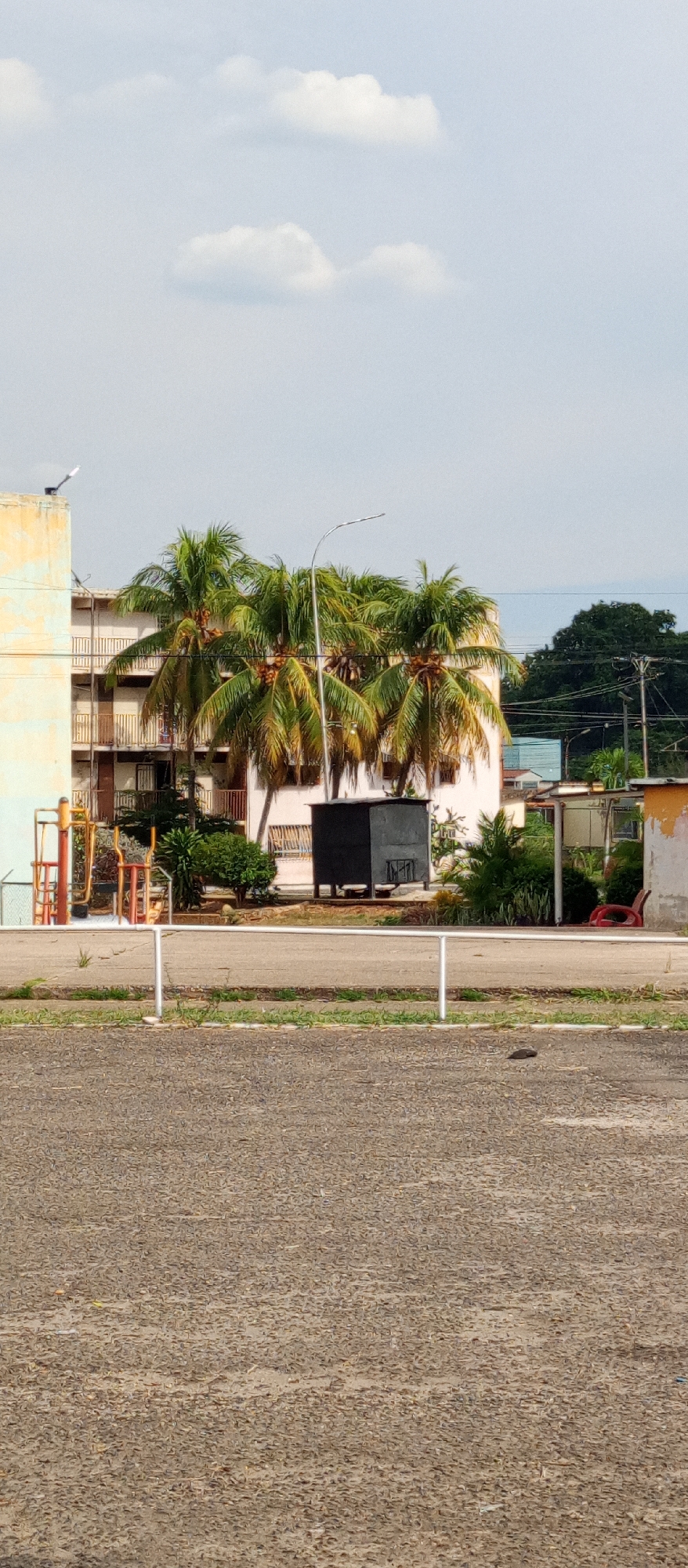 The image captures a street scene in Tinaquillo, Venezuela. The foreground is dominated by an asphalt road. A low white barrier runs across the midground, separating the road from a grassy strip. Behind the barrier, a variety of elements are visible: playground equipment, palm trees, a small dark-colored structure, and parts of several buildings, including a multi-story apartment complex in the background and single story structure to the right.. A utility pole rises beyond the buildings. The sky is overcast, with patches of blue.