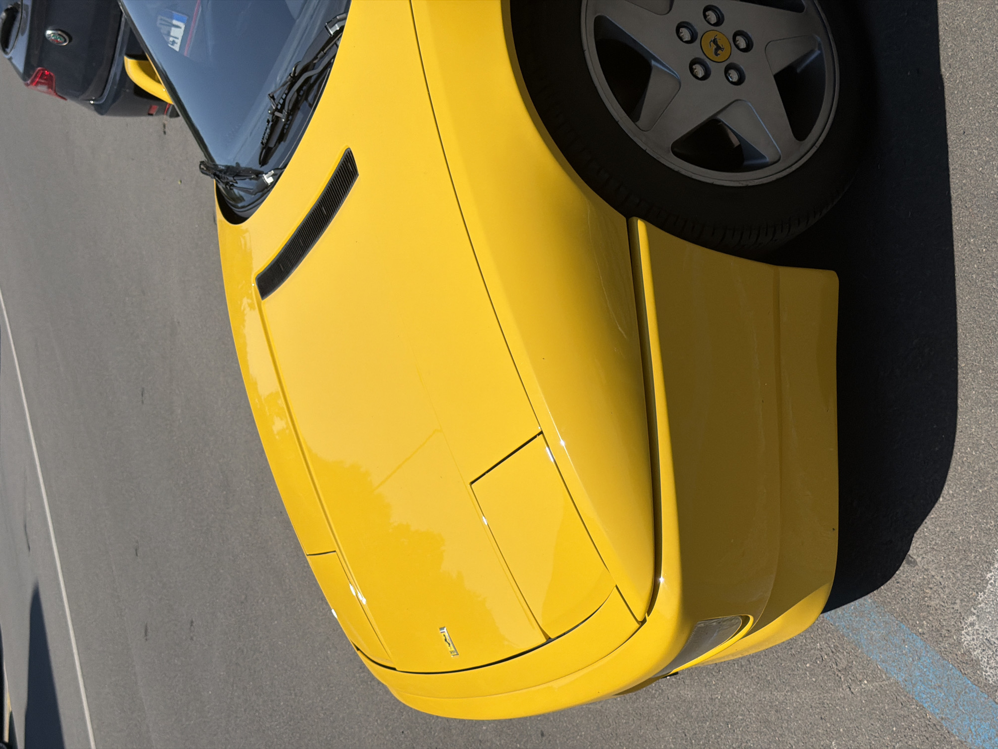 A close-up shot of the front of a yellow Ferrari car parked on asphalt in a parking lot. The angle is from slightly above and to the side, focusing on the car's hood, front left wheel, and bumper. The car's emblem is visible on the hood. The wheel has a silver rim with a black tire. The background shows a fragment of a gray car. The scene lacks any indication of being located in Milan, Italy.