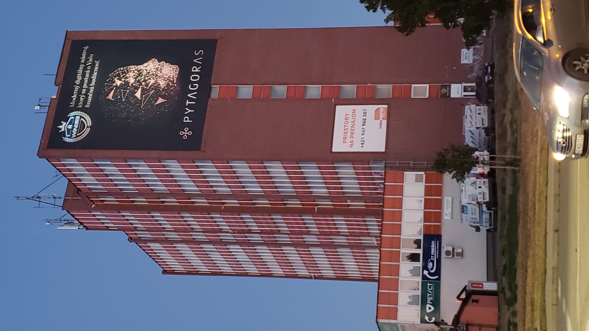 The image shows a tall, multi-story building with a reddish-brown facade, adorned with vertical bands of white and red windows. A large billboard on the upper portion of the building displays an advertisement for "PYTAGORAS" and includes a logo with a crown and the word "WINNER." The billboard's text reads: "Moderný digitálny nástroj, ktorý sa postará o Vašu finančnú budúcnosť." Below this, there is another smaller advertisement with the text "PRIESTORY NA PRENÁJOM" and a phone number. The building appears to be in an urban setting, as evidenced by the road in the foreground with vehicles, including a silver car with license plate "KE 001CH," and other structures visible to the left and lower part of the image. Signs for "PET/CT" and "CT PREŠOV" are visible on a lower section of the building, indicating a medical facility. Multiple white vans are parked in front of the building. The sky is a clear blue, suggesting it is daytime. The presence of "CT PREŠOV" suggests the location is the city of Prešov, Slovakia.