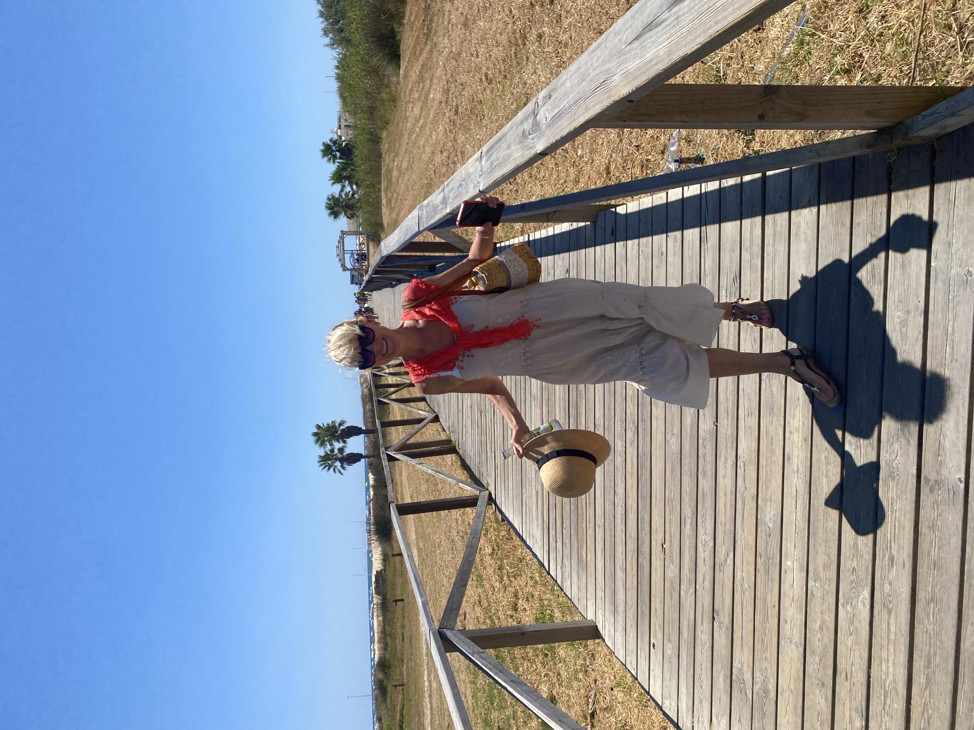 A woman walks on a wooden boardwalk toward the camera, holding a straw hat in her left hand and a smartphone in her right. She wears sunglasses, a white dress with a red scarf draped over her shoulders, and sandals. Her straw bag hangs from her left shoulder. The boardwalk, flanked by a wooden railing, is surrounded by dry grass and sparse vegetation, with palm trees visible in the background. The scene appears to be located in Conil de la Frontera, Spain, likely near a beach, as suggested by the presence of beach umbrellas and buildings in the distance. The sky is clear and blue, indicating a sunny day.