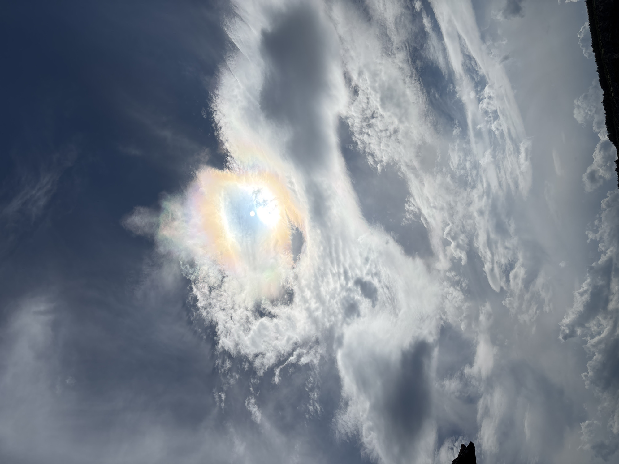 The media file captures a dramatic sky scene with a prominent sundog phenomenon. The sun is partially obscured by a cluster of clouds, creating a bright, prismatic halo around it. The sundog itself displays a vibrant spectrum of colors, including yellow, orange, pink, and blue. Wispy, streaky clouds are scattered across a deep blue sky, with larger, more opaque clouds dominating the upper and right portions of the frame. The overall impression is one of a dynamic and somewhat ethereal atmosphere. The image's orientation suggests it might be a vertical shot, emphasizing the height of the sky. There is a dark, silhouetted object in the lower right corner, possibly a part of a building or natural structure, which adds a sense of scale. The lighting suggests it is daytime, with the sun high enough to create this atmospheric optical effect. The weather appears to be partly cloudy, with conditions that favor the formation of sundogs. No people or discernible objects other than the natural elements of the sky and the silhouetted foreground are visible. There is no visible text in the image. The scene evokes a sense of wonder and natural beauty.