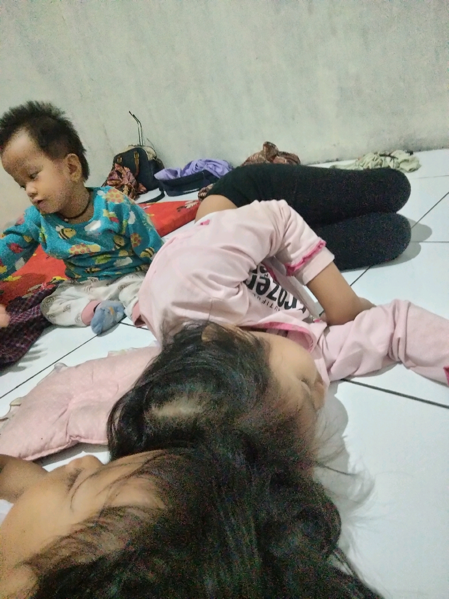 Two young children are lying on the floor in what appears to be a bedroom in Suka Sari, Indonesia. The child on the left is wearing a blue shirt with a bee pattern and white pants with a gray and white checkered pattern, and appears to be looking down. The child on the right is wearing a pink long-sleeved shirt and black pants, and is lying on their back with their head turned to the side, appearing to be asleep. A pink pillow is positioned under the head of the child on the right. Other items visible in the background include a dark bag, some folded clothing, and a red and white patterned mat. The floor is tiled with a light color. The wall in the background is textured and light gray.