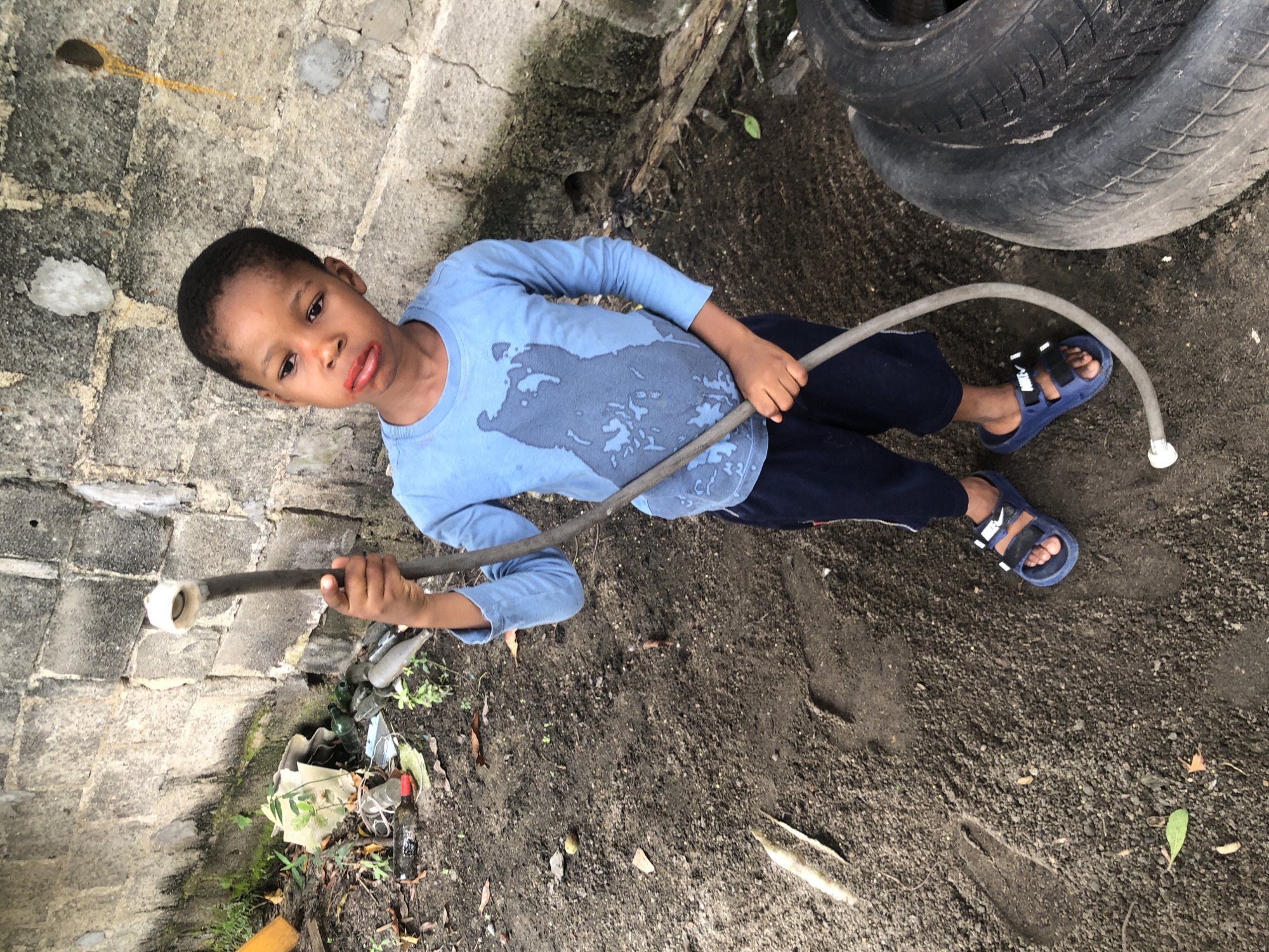A young Black boy stands in a dirt yard in Ado, Nigeria, holding a gray hose in his right hand. The boy is wearing a blue long-sleeved shirt with a dark bear graphic and navy blue shorts with blue and black sandals. The boy's lips are red, suggesting lip balm or lipstick. In the background, a rough concrete wall shows signs of wear and tear, with some green moss. Scattered on the ground are various debris, including broken glass bottles, plastic, and leaves, indicating a potentially unkempt environment. To the right of the boy, part of a large black tire is visible. The ground is covered in dark soil with footprints and scattered debris.