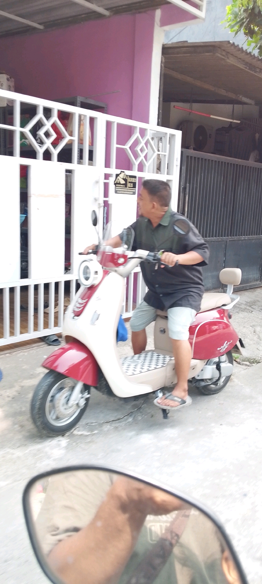 A middle-aged East Asian man in a dark short-sleeved collared shirt and light-colored shorts rides a red and cream-colored scooter. The scooter has a checkered seat pattern and a small tan passenger backrest. The man is looking to his right as he operates the scooter. He is wearing dark flip-flops. The scooter is parked on a concrete surface. In the background, there is a white metal fence with diamond-shaped openings, a pink wall, and a dark metal gate. The image appears to have been taken in the city of Satriamekar, Indonesia, likely in a residential area. A reflection of the man's legs and part of the scooter is visible in the scooter's side mirror.
