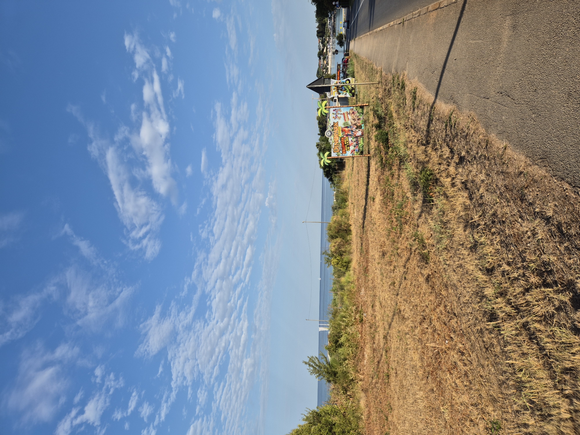 This is a landscape photograph taken in Zambratija, Croatia. The image captures a coastal scene with a road in the foreground, leading towards the sea. To the left of the road is a dry, grassy field with some scattered bushes. Further in the distance, the calm sea is visible under a bright blue sky with scattered, wispy clouds. A few sailboats can be seen on the water. On the right side of the road, a sign for "DINO PARK" is prominently displayed, featuring cartoonish characters and illustrations of dinosaurs, along with palm tree decorations. Another, smaller sign with text is partially visible to its right. Beyond the sign, there are some trees and buildings, suggesting a populated area near the coast. The overall lighting suggests it is either early morning or late afternoon.