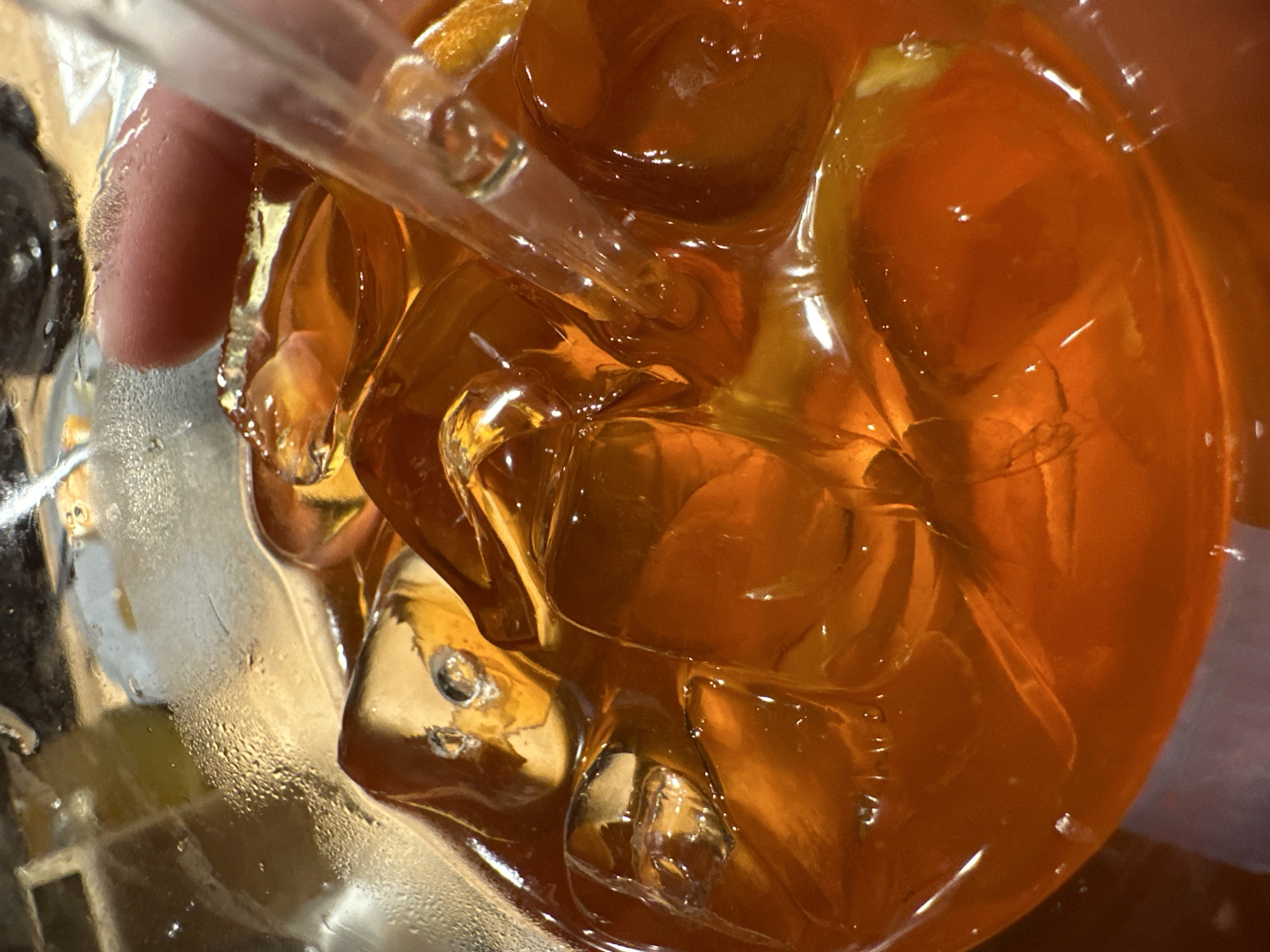 The image shows a close-up view of a glass filled with ice and a dark amber liquid, possibly iced tea or a similar beverage. A clear straw is visible, partially submerged in the liquid. The exterior of the glass has condensation droplets, indicating it is a cold drink. A finger is visible holding the straw. The image does not depict any information pertaining to Chaikovsky, Russia.