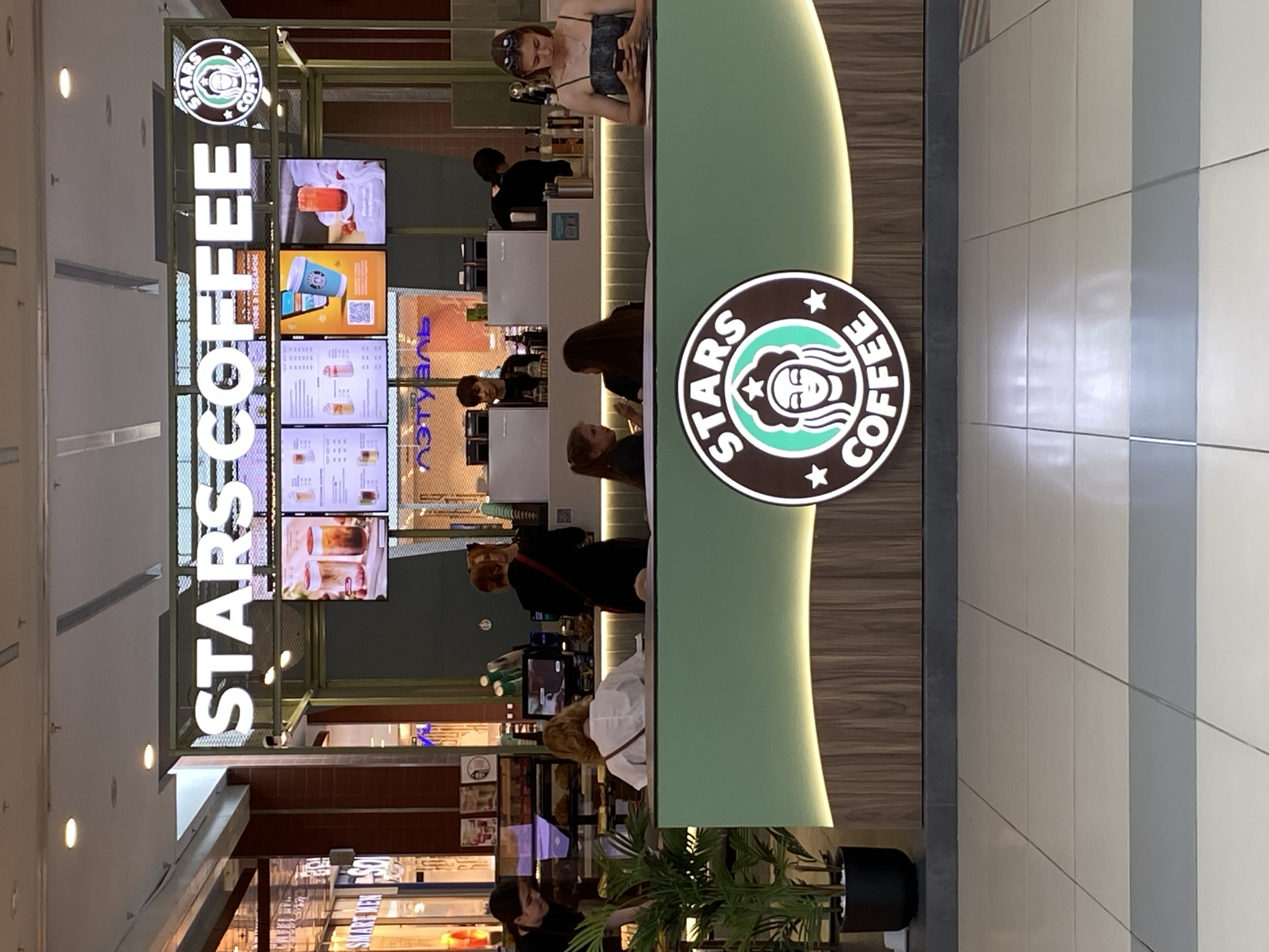 The image shows the interior of a "Stars Coffee" shop, likely a franchise, with a modern, clean aesthetic. The dominant colors are light green, white, and wood tones. Several people are visible, mostly employees and customers interacting at the counter.
In the foreground, a large circular logo for "Stars Coffee" is prominently displayed. It features a stylized face within a green circle, surrounded by stars and text. To the left, a large illuminated sign spells out "STARS COFFEE" vertically in white block letters. Behind the counter, multiple digital screens display menu items and promotional images of drinks.
Several individuals are positioned behind the counter, appearing to be employees taking orders or preparing drinks. Customers are seen interacting with them, some looking at the menu screens. The overall atmosphere appears to be a typical day at a coffee shop, with no indication of a specific event or particular time of day beyond standard business hours. The setting appears to be indoors, likely a shopping mall or a standalone commercial space. The lighting is bright and even, suggesting artificial light.