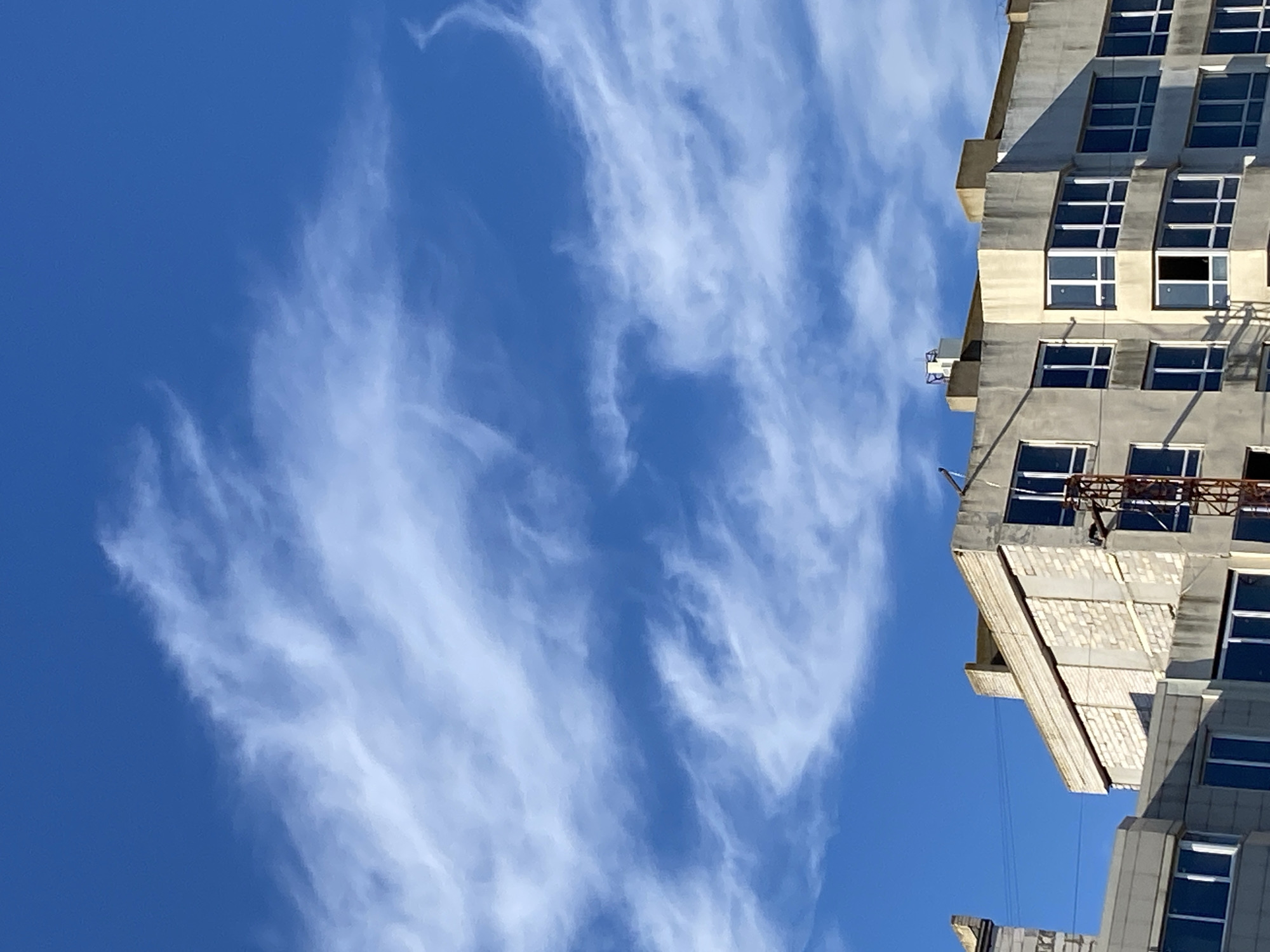 The image captures the upper floors of a multi-story building under construction against a bright blue sky filled with wispy white clouds. The building appears to be made of concrete and light-colored brick. Several windows are visible, some of which are dark, suggesting they are either tinted or reflecting the sky, while others show interior framing. A construction crane is partially visible on the right side of the frame, indicating ongoing work. The scene is set in the city of Chornomorsk, Ukraine.