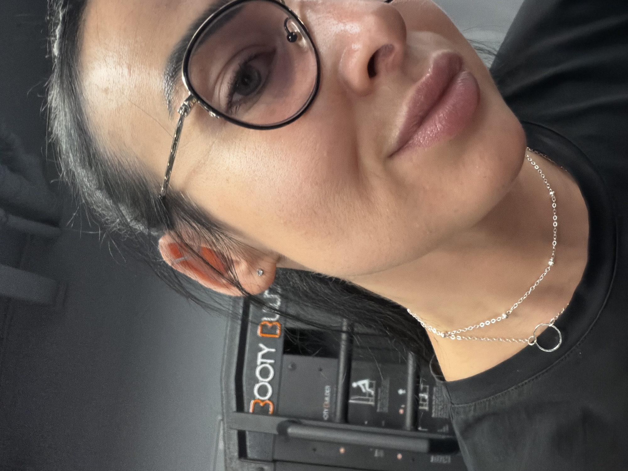 A close-up, angled shot features a person's face wearing round, black-rimmed glasses and a silver chain necklace. The person has dark hair and a fair complexion. Visible behind them is a piece of gym equipment with "BOOTY BUILDER" prominently displayed. The setting appears to be a gym or fitness facility. The city and country location is not evident from the provided image.