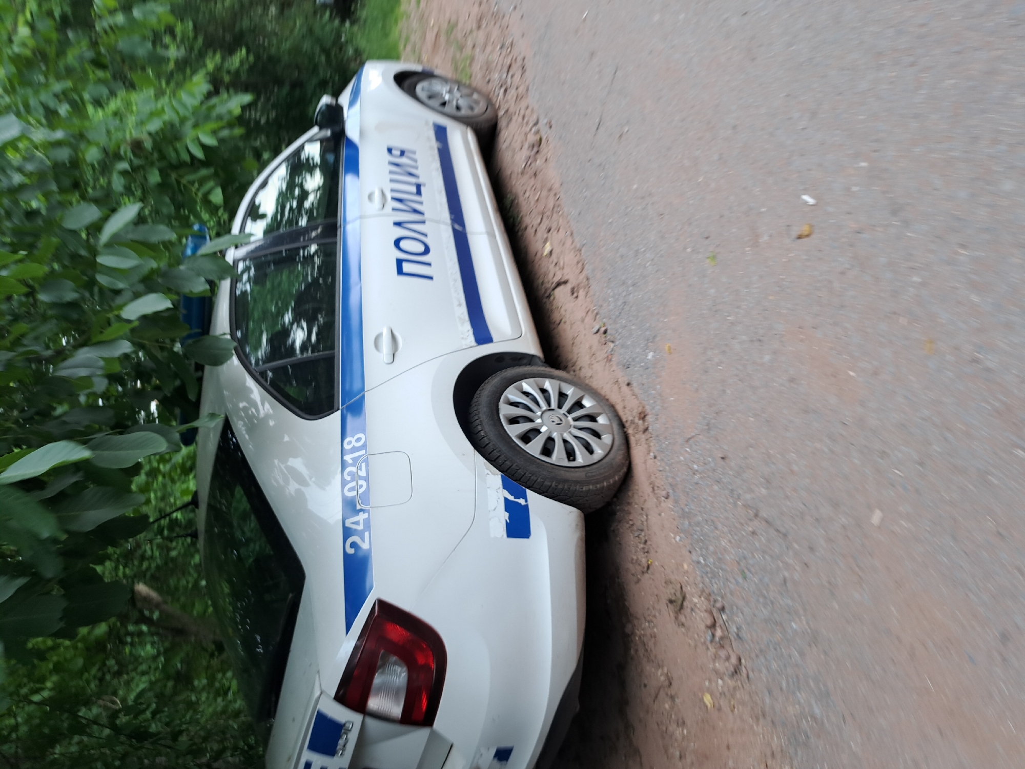 The image shows the side of a white police car parked on a dirt and asphalt road. The car has blue and white markings, with the word "ПОЛИЦИЯ" (POLITSIYA) written in blue letters on the side. There is also a blue stripe running along the length of the car. The number "24-0218" is visible in blue on the rear fender. The car appears to be a sedan.
The car is partially obscured by green foliage on the left side of the frame, suggesting it is parked near trees or bushes. The road surface is a mix of gravel and dirt, with some asphalt visible. The scene appears to be outdoors, possibly in a rural or semi-rural area, given the presence of trees and the unpaved road.
The image is taken from a low angle and is rotated, with the top of the car appearing on the right side of the frame and the bottom on the left. The lighting suggests it could be daytime, with the sun casting some reflections on the car's windows. No people are visible in the image. The overall impression is a stationary police vehicle in a natural setting.