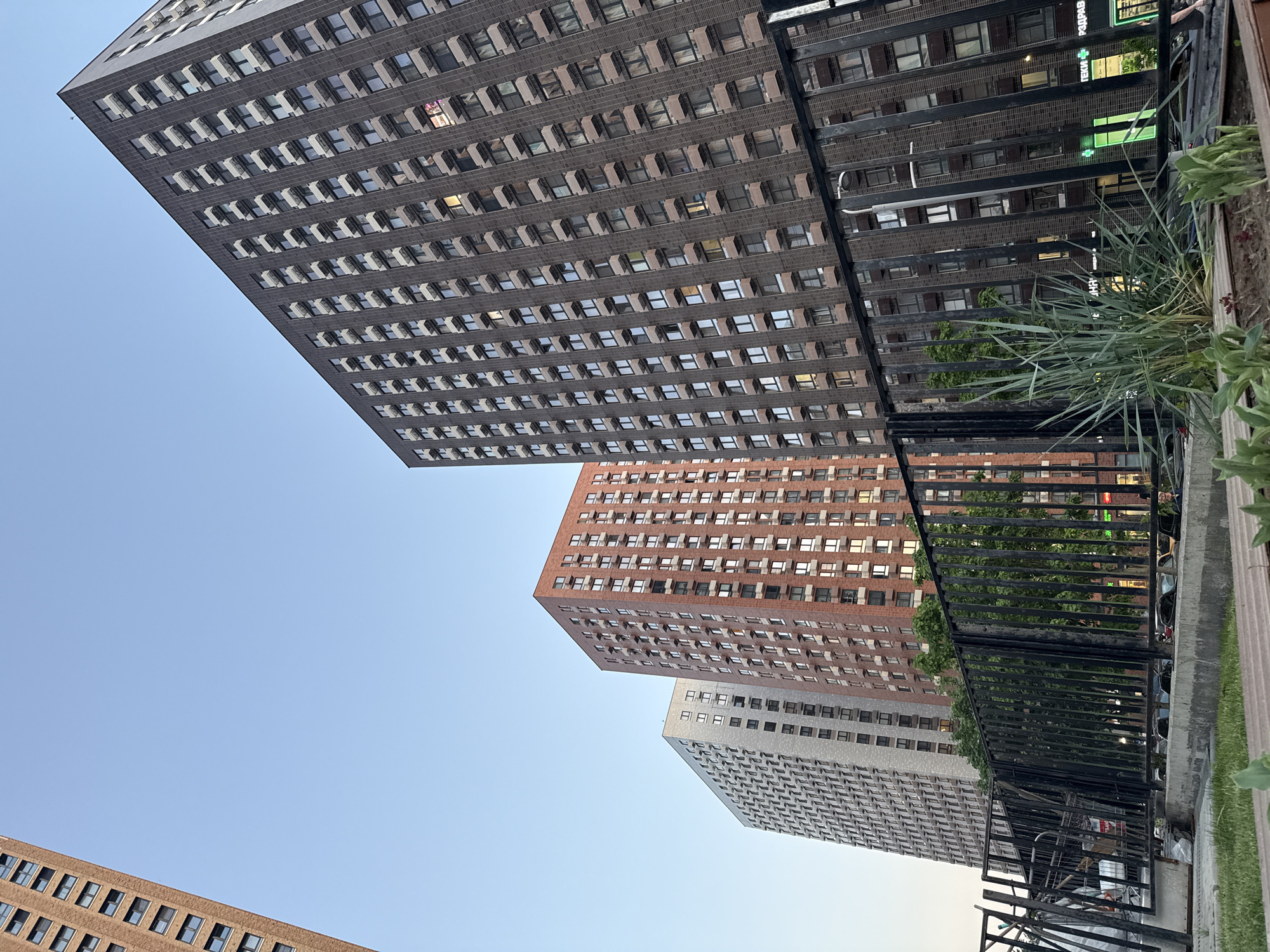 The image shows a low-angle view of several high-rise buildings in Moscow, Russia. The buildings are of different colors, one gray, one brick-red, and one tan, and feature a repetitive pattern of windows. A black metal fence and some greenery are in the foreground on the right side of the image, obscuring part of the buildings. The sky is visible in the background.