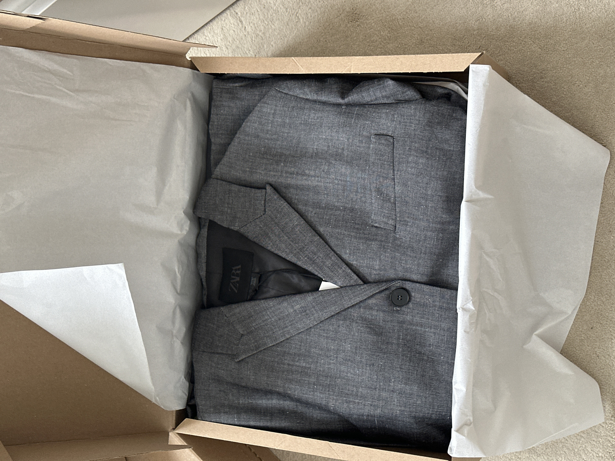 An overhead medium shot captures a grey Zara blazer neatly folded and placed inside an open, light brown cardboard box. The blazer is wrapped in white tissue paper, with a corner of the paper extending out of the box. A black Zara label is visible on the inside of the blazer. The box is resting on a light brown, textured carpet. The scene appears to be indoors, in a home environment.