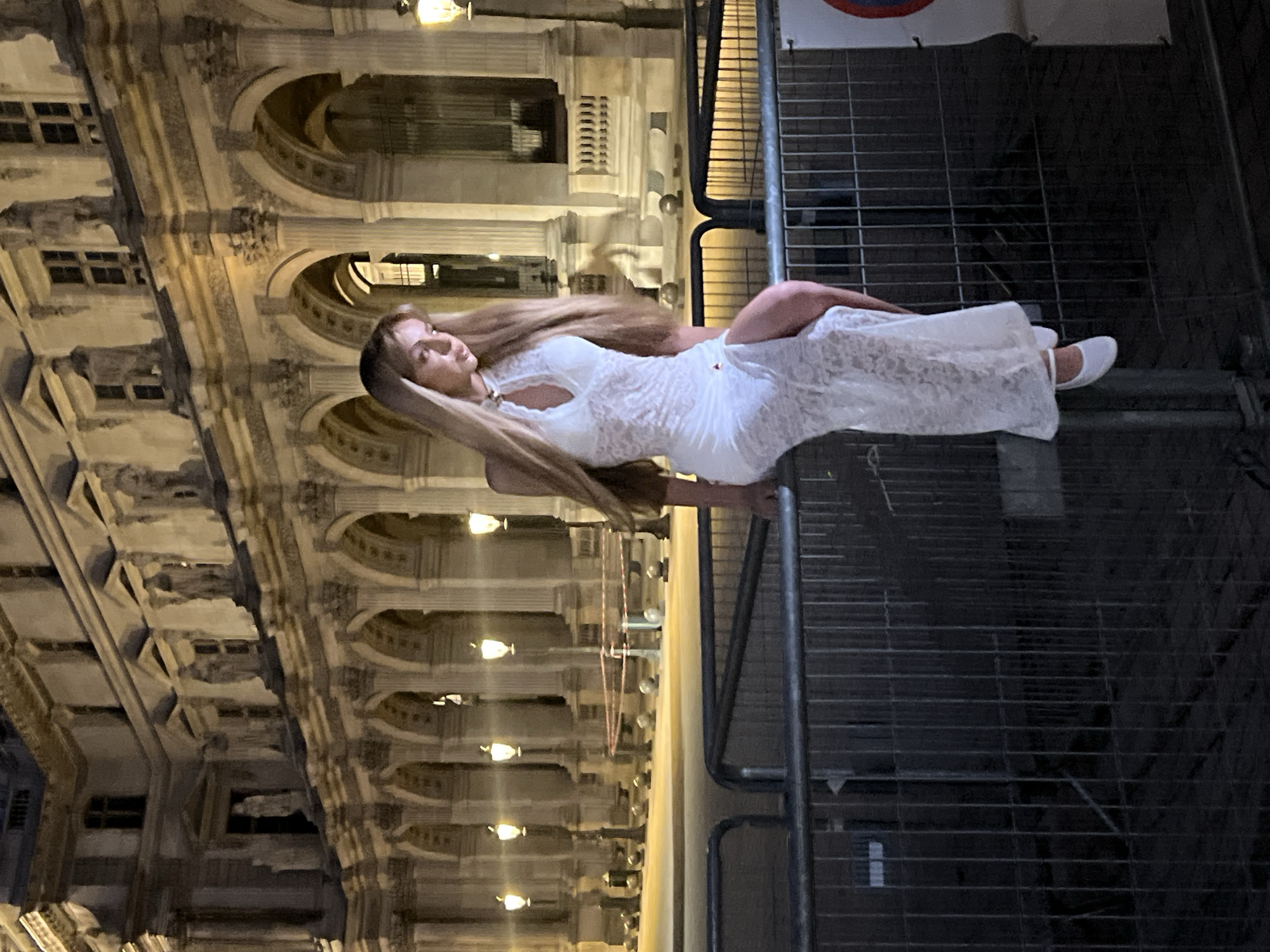 A young woman with long blonde hair is sitting on a metal barrier at night. She is wearing a white lace dress with a high neckline and a slit on the side, and white ballet flats. She is looking down and to her right. Behind her, a large, ornate building with arches and sculptures is illuminated by warm lights. The building appears to be a historical structure, possibly a museum or a palace, consistent with the architecture found in Paris, France. The scene is framed by a metal barrier with a red and white sign partially visible on the right side. The overall atmosphere is elegant and nocturnal.