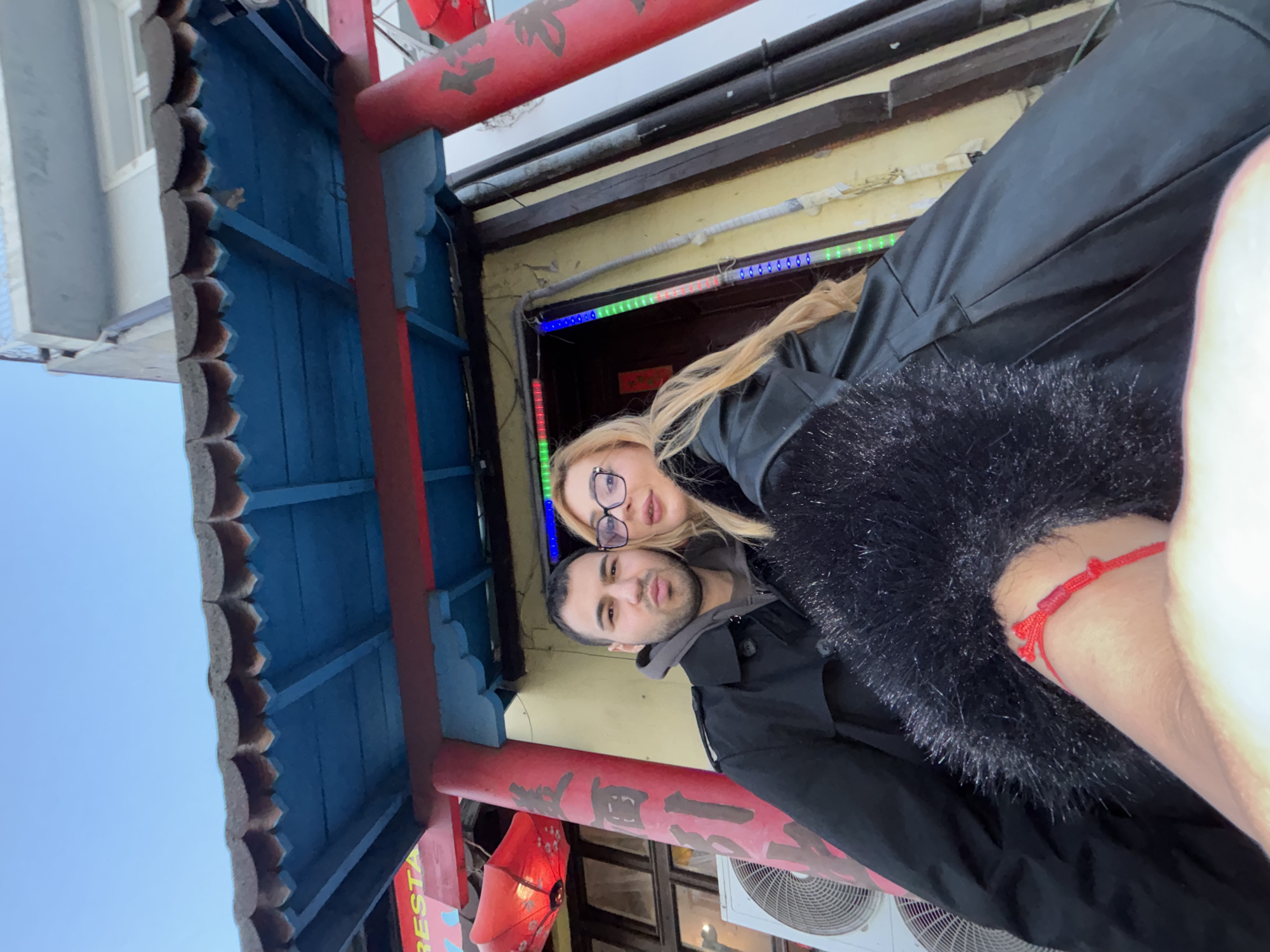 A selfie of a man and a woman, likely taken by the man, standing under the ornate entrance to a building, perhaps a restaurant or store, in Varna, Bulgaria. The man has dark hair and a slight beard. The woman has blonde hair and wears glasses. They are both wearing dark jackets. The entrance is adorned with red and blue decorations, and Chinese characters. There is a red lantern visible. The architectural style is reminiscent of a traditional Chinese entrance. In the background, there are windows and an air conditioning unit. A red bracelet on the wrist is visible at the rightmost edge of the image.