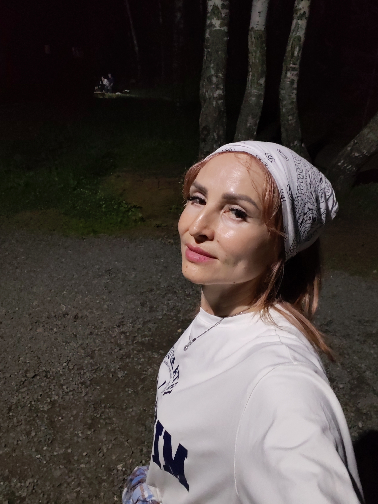 A woman with light brown hair, wearing a white bandana and a white t-shirt, is standing outdoors at night. She is looking towards the camera and smiling. The background shows several birch trees and dark foliage. In the distance, a group of people are gathered around a light source, possibly a campfire. The ground appears to be covered in gravel. The location is identified as Chaikovsky, Russia.