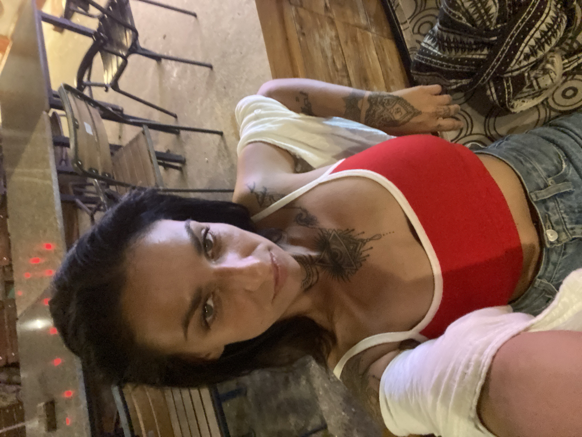 A woman with dark hair and tattoos is taking a selfie in what appears to be an outdoor seating area of a restaurant or bar. She is wearing a red tank top with white trim and blue jeans. A white garment is draped over her shoulders. The background shows tables and chairs, with some red lights visible on a surface in the distance. The floor is concrete. The image is taken from a high angle.
