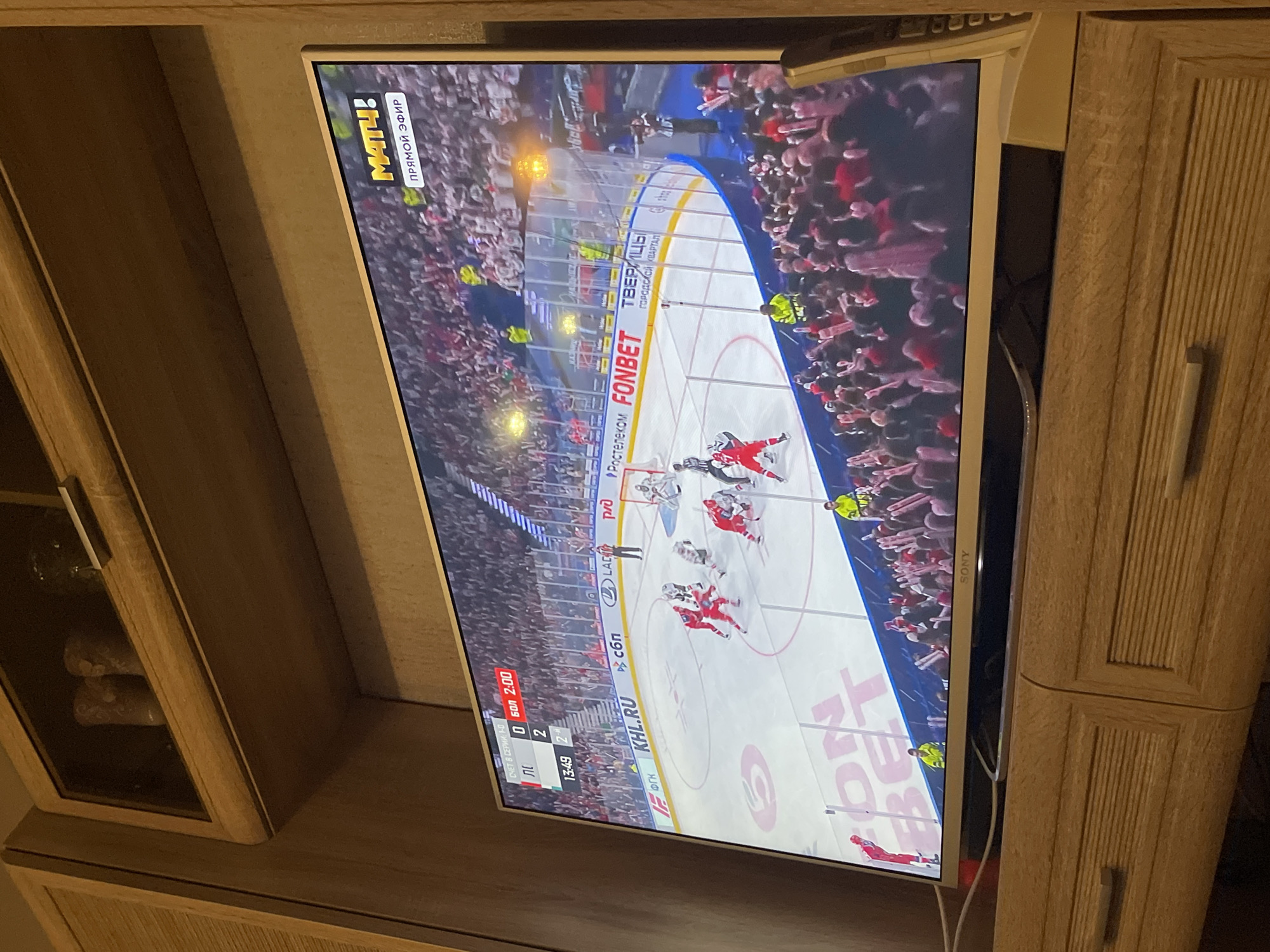 A medium shot shows a television, which is presumably a Sony television based on the logo visible on the right edge of the screen, airing a live hockey game. The hockey teams wear red and black uniforms. The television is situated in a room with furniture in natural oak. The hockey match is sponsored by companies such as FONBET and Rostelecom. The broadcast channel is "Match TV," identified by the logo on the top-left of the screen, which is airing "Прямой Эфир" or "Live" as translated. According to the screen, the time in the game is 13:49 in the second period, and the score is 0-2. The location "ТВЕРИЦЫ ГОРОДСКОЙ КВАРТАЛ" is displayed on the ice rink. Though Tveritsy is in Russia, it does not appear to be in the city of Перевозное.
