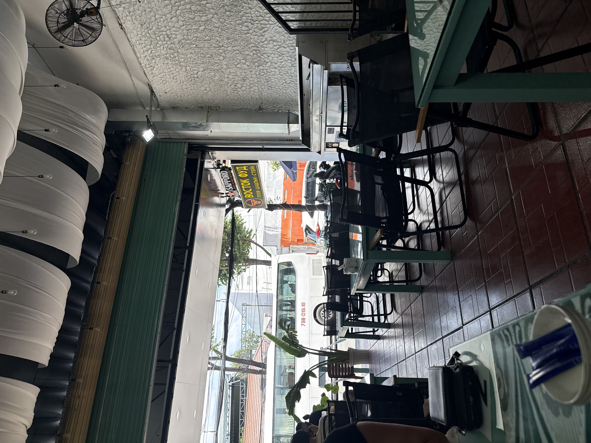 This is an interior view of a restaurant in Nha Trang, Vietnam, looking out through an open storefront. The restaurant has a rustic aesthetic with a tiled floor and green metal tables and chairs. A large plant is situated near the entrance. Outside, a white bus is parked on the street, and there are trees and buildings visible. A sign in Cyrillic script, likely indicating the restaurant's name and offerings like "plov, shashlik, steak," is prominently displayed near the entrance. The ceiling is adorned with draped white fabric, and several light bulbs hang from it. A ceiling fan is visible in the upper right corner. The scene appears to be daytime.