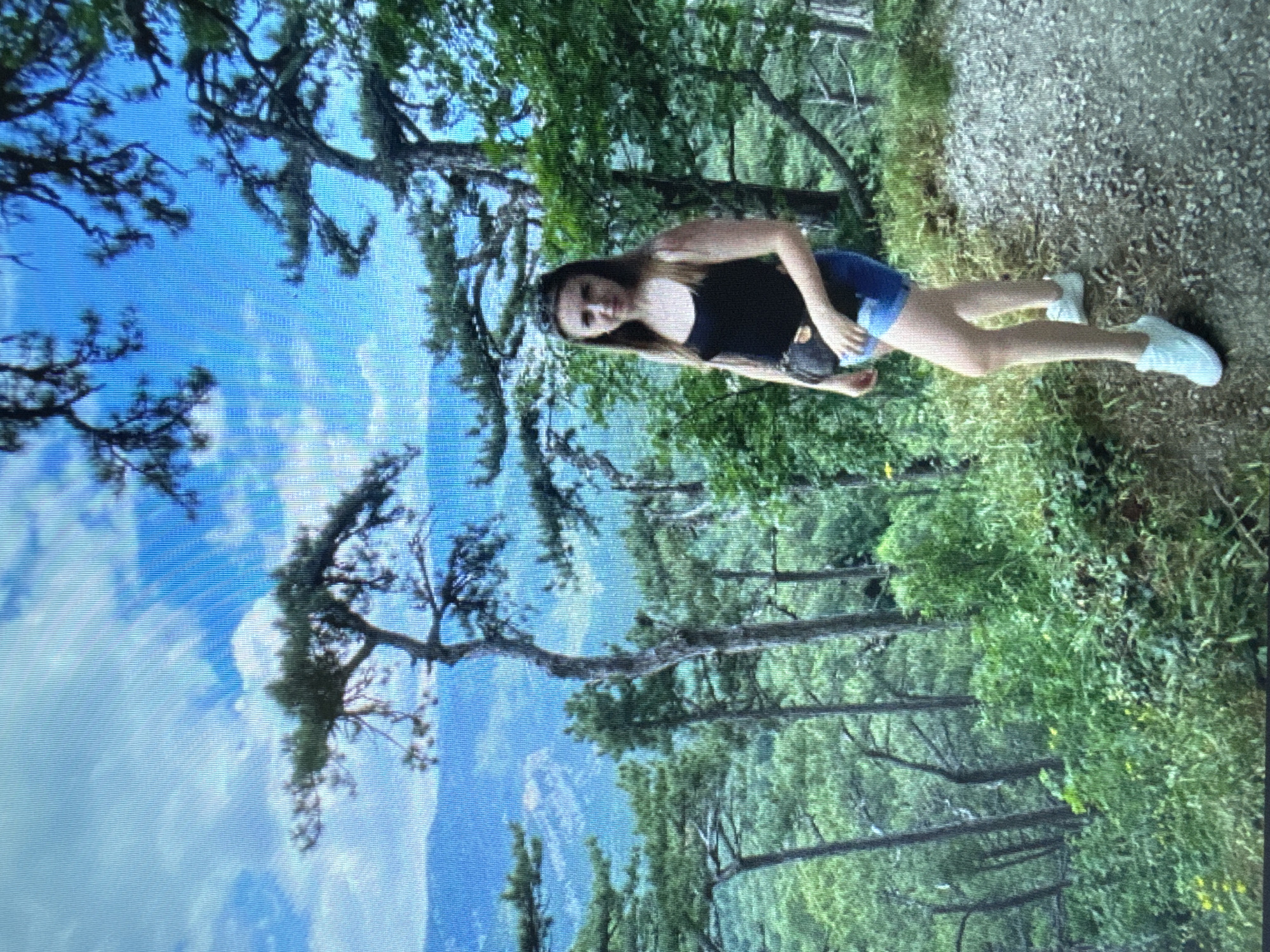 A young woman with long brown hair stands on a gravel path in a forest. She is wearing a black tank top, blue denim shorts, and white sneakers. A small black bag is slung across her body. The path is to the right of the frame, and a dense forest with tall trees and lush green foliage is to the left. In the background, mountains are visible under a bright blue sky with scattered clouds. The lighting suggests it is daytime.