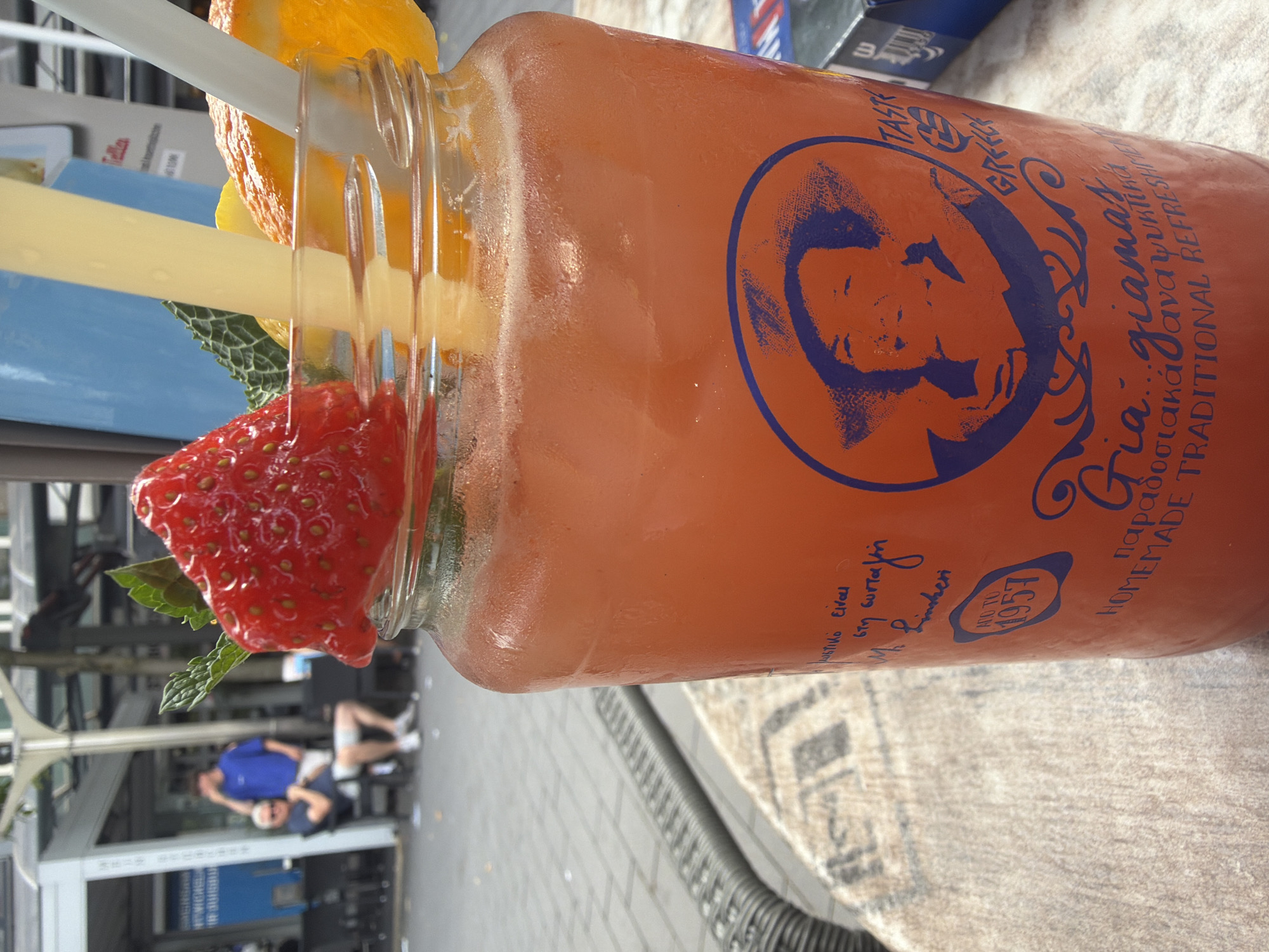 The image shows a close-up of a glass jar filled with a red beverage, likely a cocktail or juice, adorned with a strawberry, mint leaves, and slices of orange and lemon. Two straws, one yellow and one white, are inserted into the drink. The jar has a distinctive orange exterior with blue text and graphics, including a portrait and the words "Gia...giamas" and "HOMEMADE TRADITIONAL REFRESHMENT." A smaller blue label with "ESTD. 1957" is visible on the side. The drink is placed on a table with a textured surface. In the blurred background, several people are visible, some seated and others standing in an outdoor setting. The overall environment suggests a cafe or restaurant terrace. The presence of a Coca-Cola cigarette pack on the right side of the frame, and an article referencing Duisburg in the provided crops, indicates this scene is likely located in Duisburg, Germany.