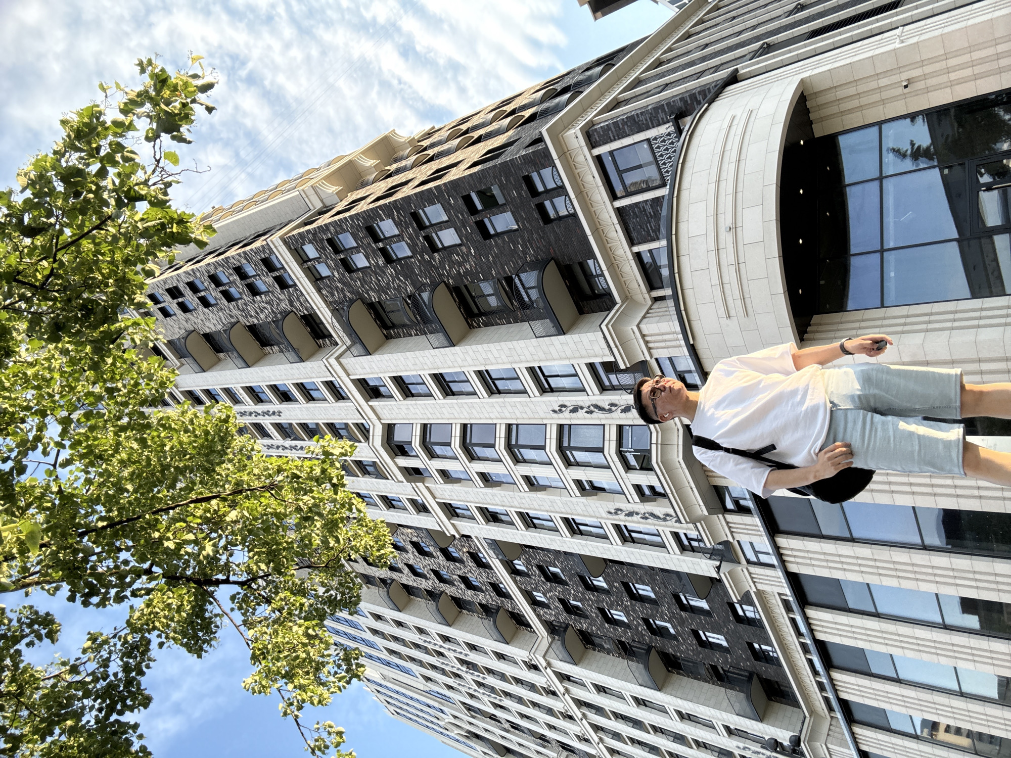 An upward-angle medium shot captures a young man standing in front of a modern, multi-story building with a dark brick facade and cream-colored decorative elements. The building features numerous windows and balconies with ornate railings. Green foliage from a tree drapes over the top left of the frame, partially obscuring the blue sky with scattered white clouds. The man, wearing a white t-shirt, light blue shorts, glasses, and earbuds, has a black backpack slung over his right shoulder. He is looking to his right, with his left hand resting on his thigh. The scene appears to be in Moscow, Russia, based on architectural style and common building materials found in the city.
