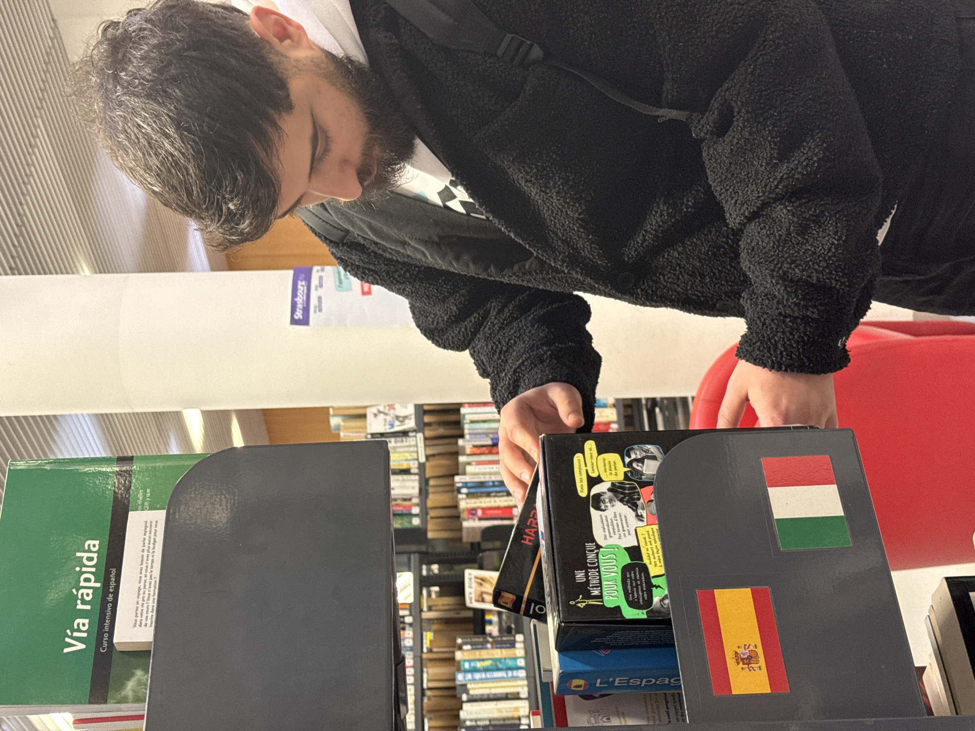 A bearded person wearing a black fleece jacket is looking at books on a library shelf. The books are mostly language learning materials. On one book, the flags of Hungary and Spain are displayed. "Vía rápida" is visible on one of the green books. A sign "Strasbourg" is on a white pillar in the background.