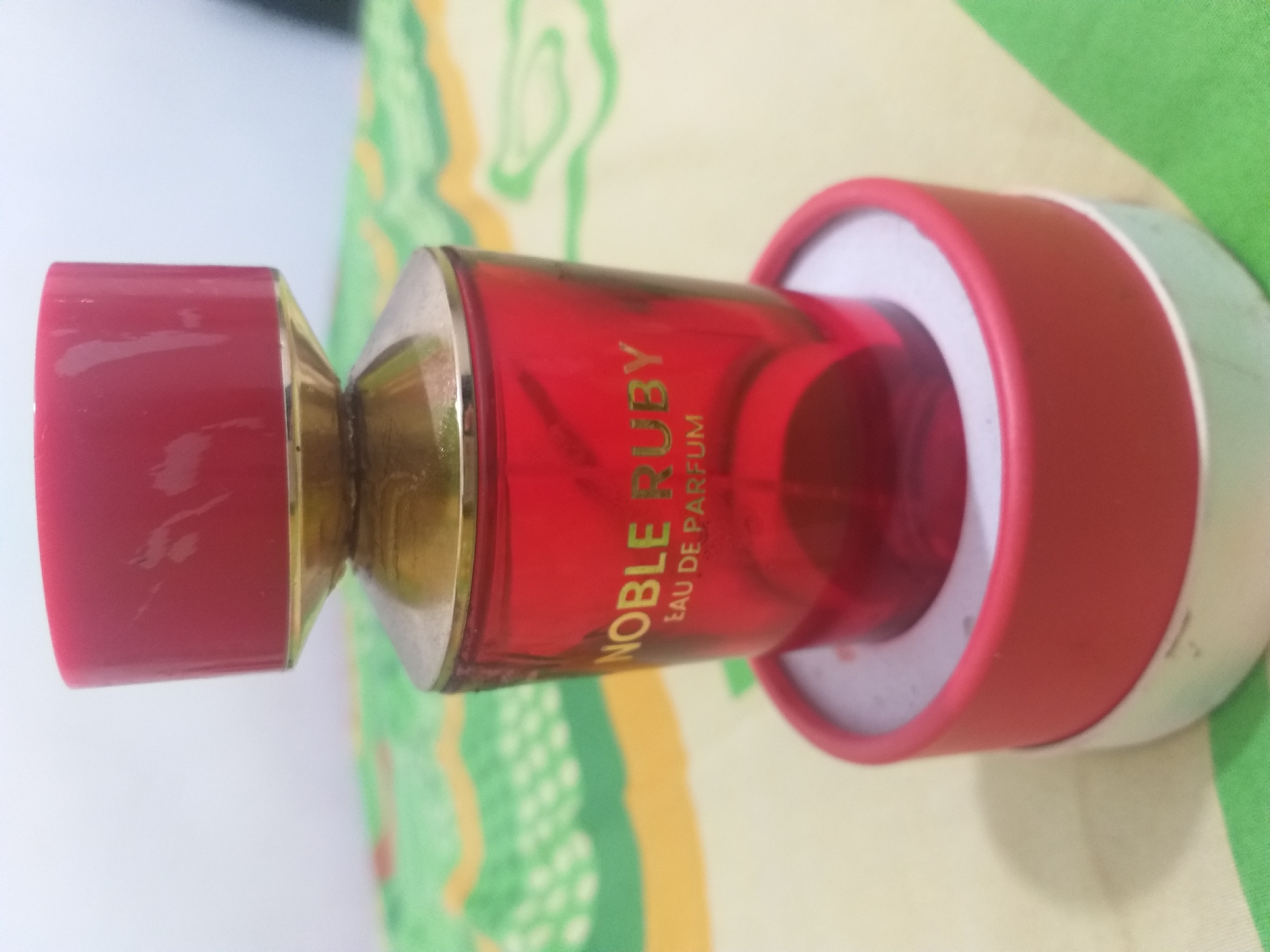 A close-up, eye-level shot shows a red glass perfume bottle labeled "NOBLE RUBY EAU DE PARFUM" in gold lettering. The bottle has a gold rim and a red cap. It sits on a red circular base, which in turn rests on a colorful, patterned surface that appears to be a tablecloth. The background is softly blurred. No people or animals are visible. The setting is likely indoors. The image does not provide any information about the city of Air Jamban, Indonesia.