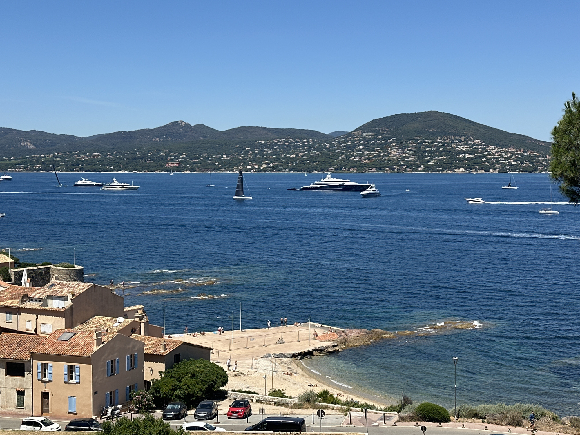 This is a high-angle, wide shot of the coast of Saint-Tropez, France, on a sunny day. The scene is dominated by a vast expanse of deep blue water, reflecting the clear blue sky. In the foreground, the terracotta roofs of several buildings characteristic of the Mediterranean architectural style are visible. These buildings appear to be part of a town or village, with windows and shutters adding detail.
The water is dotted with numerous vessels of varying sizes. Several large yachts, some appearing luxurious and modern, are anchored or cruising on the sea. Smaller sailboats, including at least one with a black sail, are also present, indicating recreational sailing activity. In the distance, the coastline is lined with lush green hills, covered in trees and dotted with numerous houses, suggesting a densely populated residential area. The hills rise gently, forming a picturesque backdrop to the seascape.
Closer to the shore, a concrete pier extends into the water, with a small sandy beach adjacent to it. Several people can be seen on the pier and the beach, some sunbathing, others standing or walking. There are also some scattered rocks along the coastline. The presence of parked cars on a road in the lower part of the image indicates that the area is accessible by road. The overall impression is one of a vibrant coastal resort town enjoying pleasant weather, with a mix of residential areas, leisure activities, and maritime presence.