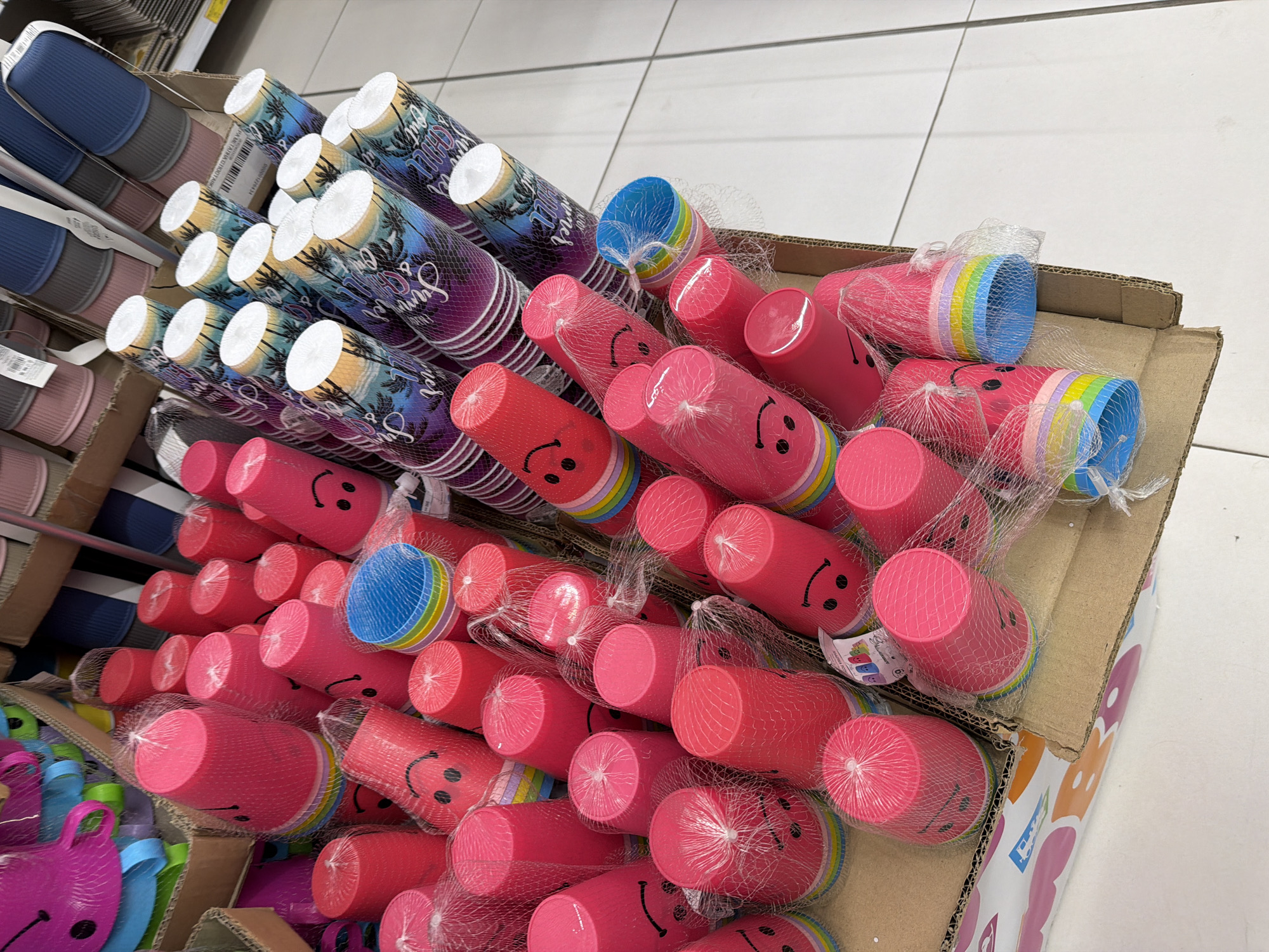 The image depicts a retail setting, likely a store, featuring an assortment of colorful plastic cups. The cups are predominantly pink and feature smiley face designs. They are packaged in netting and stacked within cardboard boxes. In the background, other items are visible, including what appear to be slippers or sandals in shades of blue, gray, and pink, also organized on shelves or in display boxes. The flooring appears to be tiled. The scene is brightly lit, suggesting an indoor commercial environment. There are no people visible in the image. The location is Plovdiv, Bulgaria.
