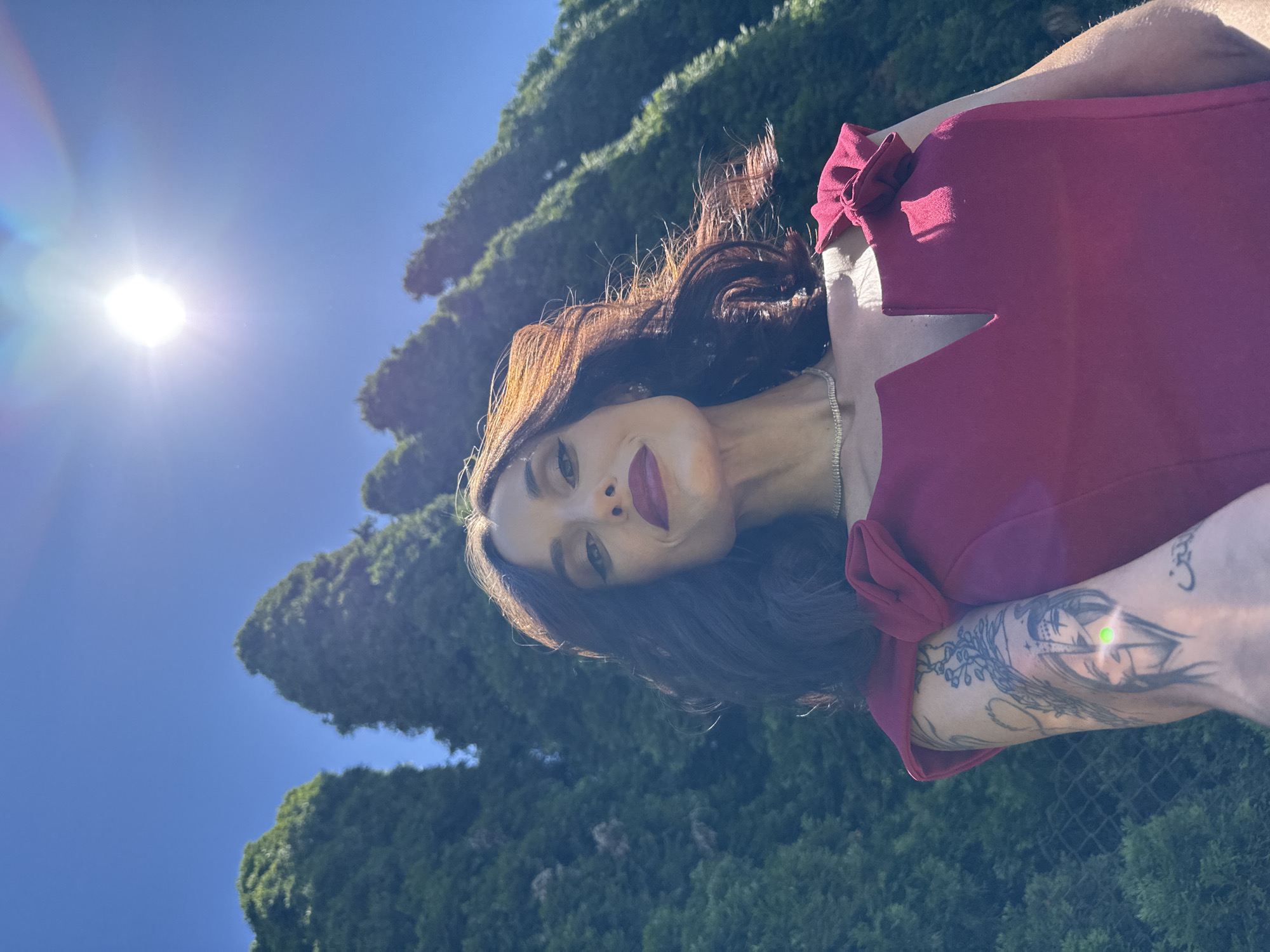 The image shows a woman with dark hair and a burgundy dress standing outside in a sunny environment with trees and a blue sky visible. The woman is fair-skinned with red lipstick and a silver necklace, and she has a tattoo on her arm. The location is not identifiable as Ribaritsa, Bulgaria, without additional context.
