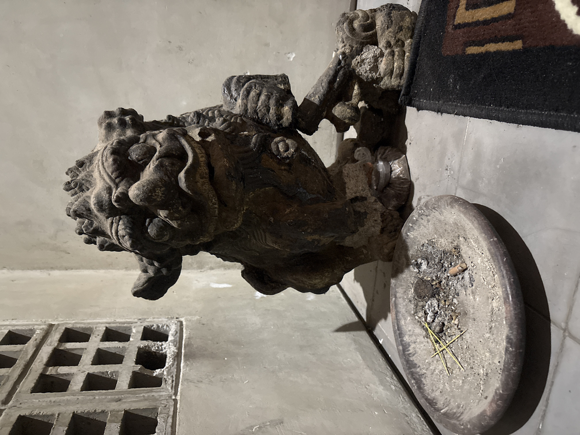 The scene is an interior view in Kalijaten, Indonesia, under low, direct lighting. A large, dark, weathered statue, sculpted from a rough material like stone or concrete, resembling a guardian lion or Foo Dog, occupies the mid-right. It features detailed facial characteristics, a prominent mane, and its right front paw rests upon a textured, spherical object. The statue casts a distinct shadow on the light gray floor. On the upper-left wall, a light gray concrete ventilation block with a grid pattern is visible; one lower segment of the block shows a chipped or broken portion. In the lower-left foreground, an oval, unglazed earthenware dish serves as an ash receptacle, containing a quantity of ash, four thin yellow spent incense sticks arranged in a cross, and a single cigarette butt. The walls are light gray, textured, and appear to be painted concrete or plaster. A section of a dark-colored woven rug, displaying brown and tan patterns, is partially visible on the floor in the lower-right. No individuals are present within the frame.