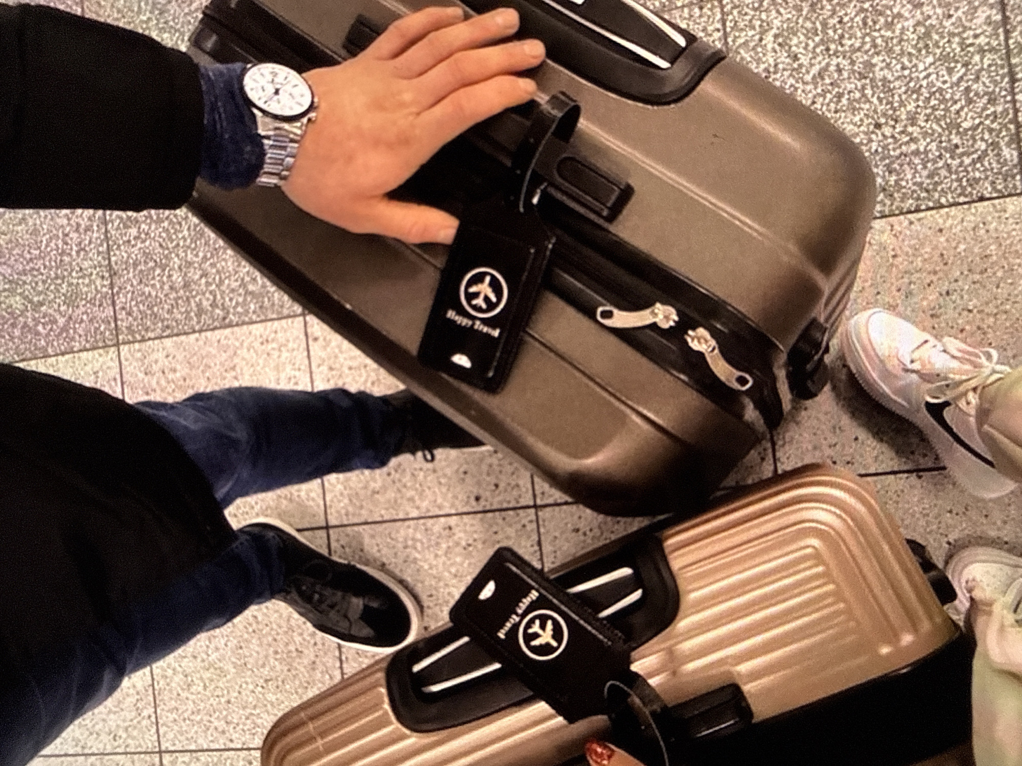 The image shows a top-down view of two people standing next to luggage on a tiled floor. One person is wearing dark jeans and black sneakers. The other person, on the right, is wearing dark pants and has a watch on their left wrist. They are both holding onto luggage. There are two suitcases visible: a gold one on the left and a bronze one on the right. Both suitcases have black luggage tags attached, each with an airplane logo and the text "Happy Travel". The background appears to be an indoor public space, possibly an airport or train station, given the presence of luggage and the tiled floor. There is no specific indication of Varna, Bulgaria.