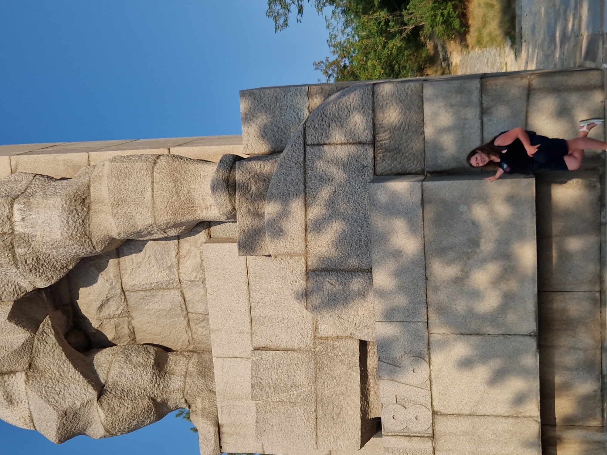 An adult female, with long, brown hair, wearing a black tank top and dark blue shorts, stands to the right of a massive stone monument in Panagyurishte, Bulgaria. She is leaning against the monument with her left hand and has her right leg bent at the knee. She is smiling at the camera. The monument is made of large, textured stone blocks and appears to be part of a larger sculpture. The lower portion of the monument has the numerals "1876" carved into it. Tree shadows are cast across the monument. In the background, there are trees and a pathway. The sky is clear and blue.