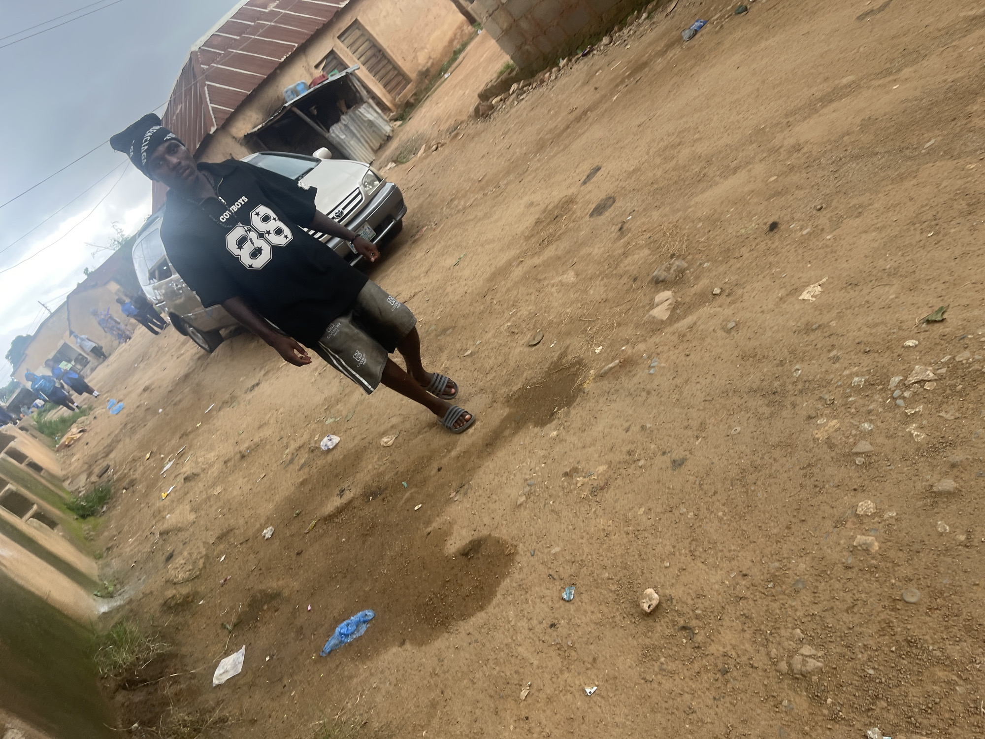 A young man, wearing a black beanie and a black t-shirt with "COWBOYS 88" printed on it, stands in a dusty outdoor area. He is also wearing gray shorts and flip-flops. The man is looking towards the camera with a neutral expression. In the background, a white car is parked, and some buildings with corrugated metal roofs are visible. The ground is covered in dirt and small stones, with some scattered debris including plastic bags. A cloudy sky suggests overcast weather. The scene appears to be in a village or rural area, with hints of local architecture and unpaved roads. Some other people are visible in the distance, appearing to be walking or standing. The overall impression is of a casual, everyday scene in a somewhat neglected urban or semi-urban environment.