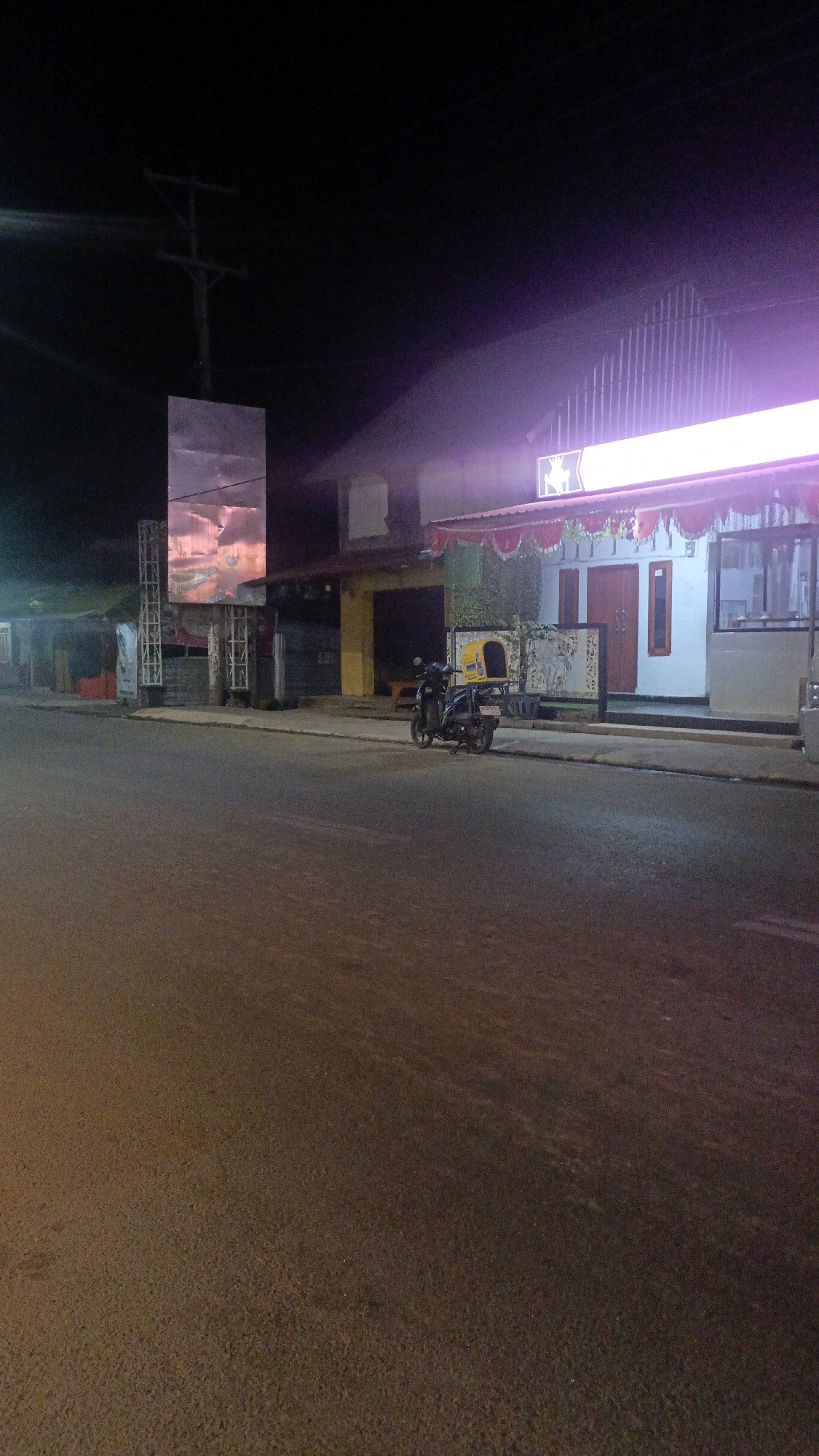 The image depicts a nighttime street scene in Taliwang, Indonesia. A paved road occupies the foreground, with a sidewalk running alongside a building on the right. A motorcycle, equipped with a yellow delivery box on the rear, is parked on the sidewalk. The building features a white facade with red-trimmed awnings and two prominent red doors. Above the building, a brightly lit sign displays a graphic of a deer. To the left of the building, a large, reflective billboard stands tall, displaying an image of a sunset or clouds. The background is dark, with a utility pole visible in the upper left quadrant. The lighting suggests artificial illumination from the building's sign and the billboard.