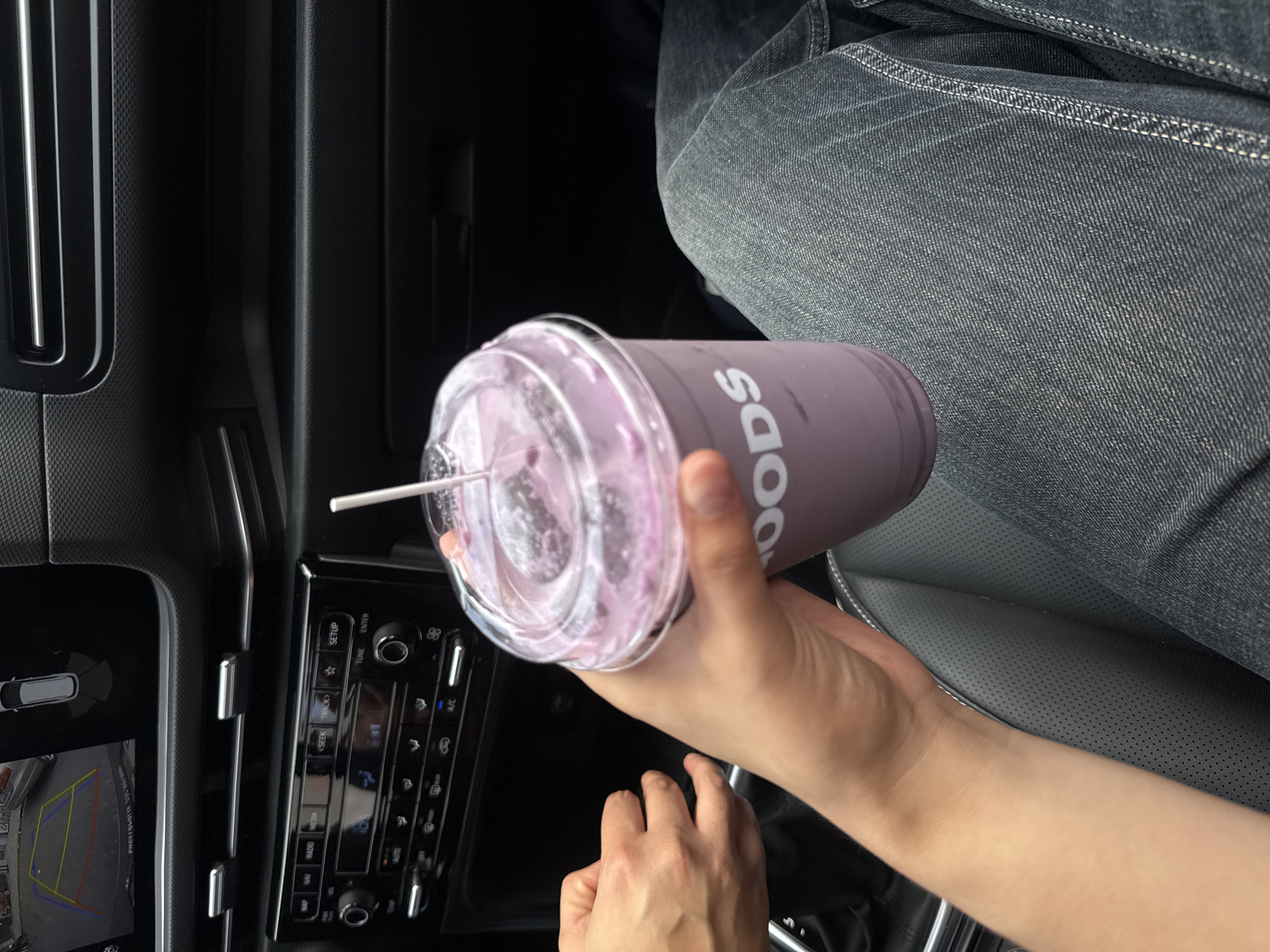 A close-up, overhead shot shows a person in Puebla, Mexico, seated in a car. They are holding a purple beverage with a straw in a clear plastic cup with the brand name "Woods" visible on the side. The car's dashboard and console are visible, including a screen showing a backup camera view. The person is wearing denim jeans. One hand holds the cup, while the other is visible further down, closer to the gear shift.