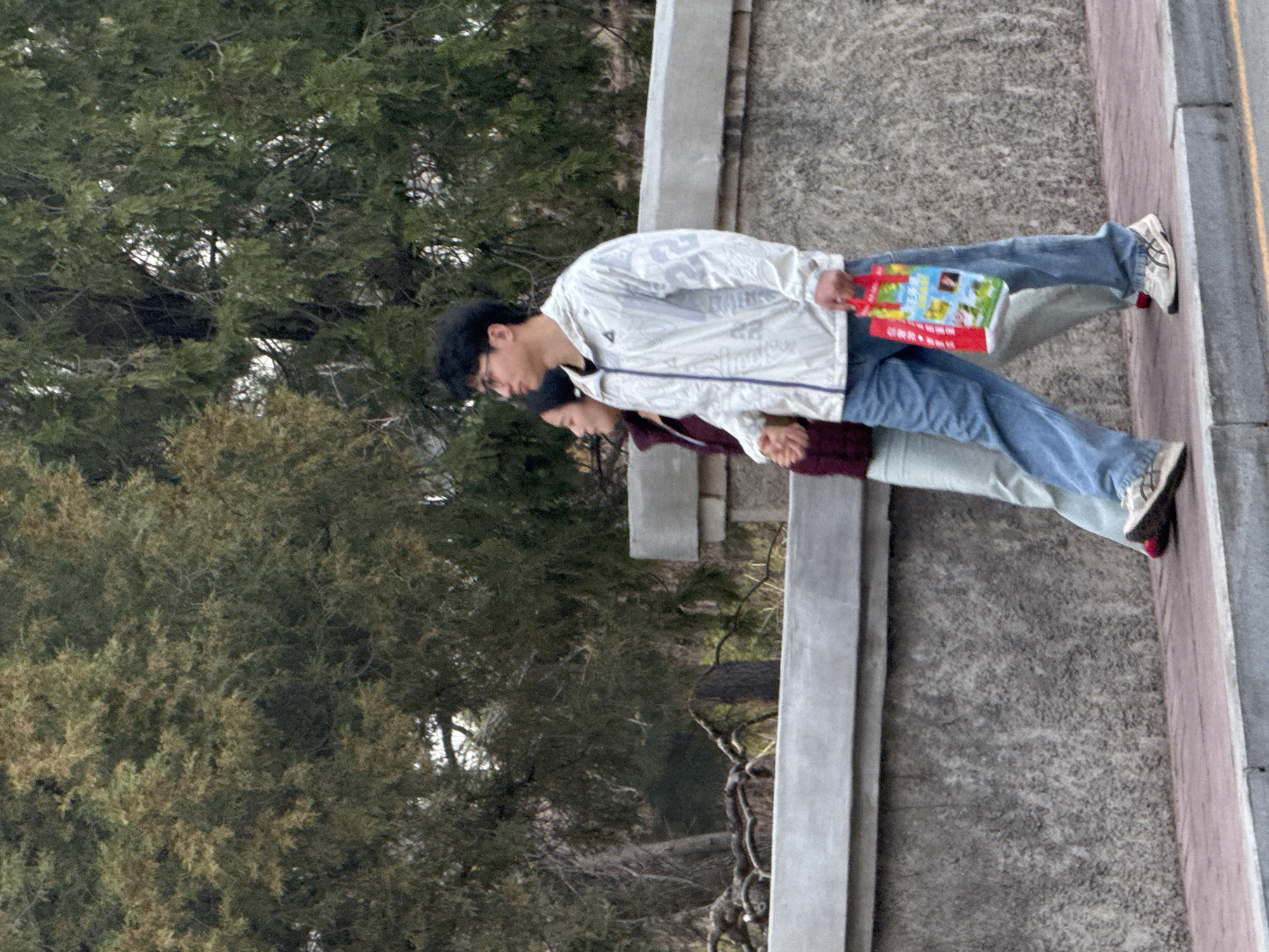 A medium shot captures a couple walking side by side on a sidewalk alongside a road in Zhongtun, China. The man on the left is wearing a white jacket with light blue jeans and white sneakers. He is carrying a book in his left hand. The woman beside him is wearing a maroon sweater with light blue jeans. The background consists of a cement barrier on the right, trees on the left, and a road in the far distance. The setting appears to be outdoors during the day.