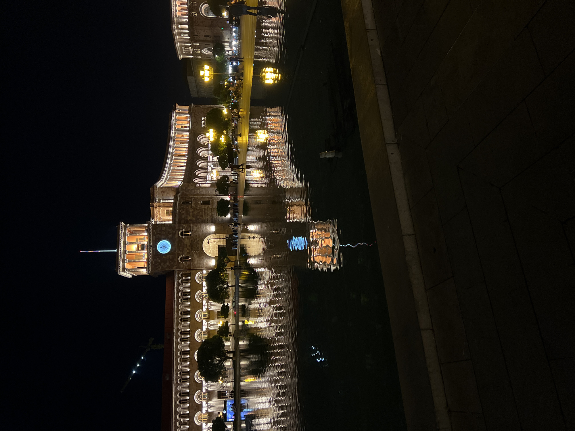 A wide-angle, nighttime shot captures the illuminated facade of a grand building with a prominent clock tower, reflecting in a dark body of water in the foreground. The building features arched windows and ornate architectural details, with warm lights spilling from within. A flag flies atop the clock tower. Several trees are visible along the building's edge, and in the distance, streetlights and car headlights illuminate the urban landscape. People are scattered throughout the scene, some walking, others seated, creating a sense of activity. The water's surface is disturbed, causing distorted reflections of the lights and the building. The sky is dark, with a hint of a construction crane in the upper left quadrant. The ground in the very foreground is paved with large, dark tiles, with a concrete border along the edge of the water.