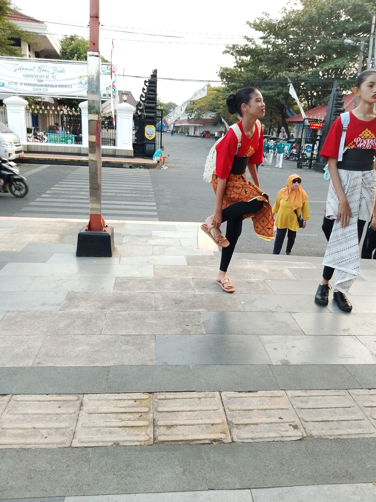 In the city of Pati, Indonesia, a group of young women in traditional attire pose on a set of stone stairs. The woman in the foreground, wearing a red shirt, black sash, and patterned skirt over black leggings, stands on one leg with her arms outstretched. To her right, another young woman in a similar red shirt and patterned skirt walks away from the camera. Further back, a woman in a yellow headscarf and dark pants is visible. The background features a street with a zebra crossing, a white banner with Indonesian text, a black gate with decorative elements, and trees. The overall scene suggests a cultural event or performance taking place outdoors.