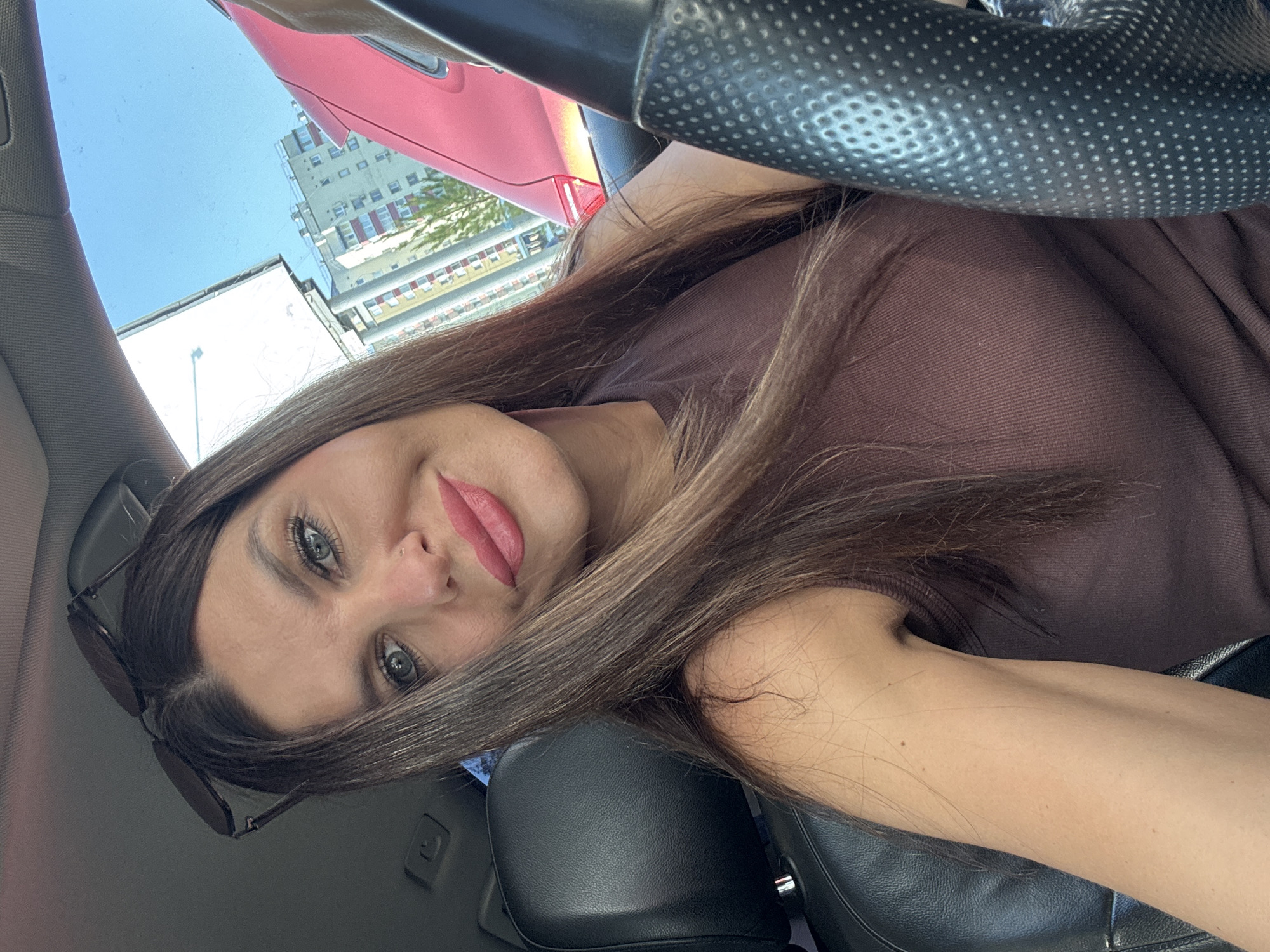The image shows a close-up selfie of a woman in a car. She has long, brown hair, blue eyes, and is wearing pink lipstick. Sunglasses are perched on top of her head. She is wearing a brown tank top. The interior of the car is visible, including a black leather seat and part of the steering wheel. Outside the car, buildings and a bright blue sky are visible, suggesting an urban environment. The presence of buildings that appear to be apartment blocks and the overall urban setting are consistent with the city of Chaikovsky, Russia. The lighting indicates it is daytime.