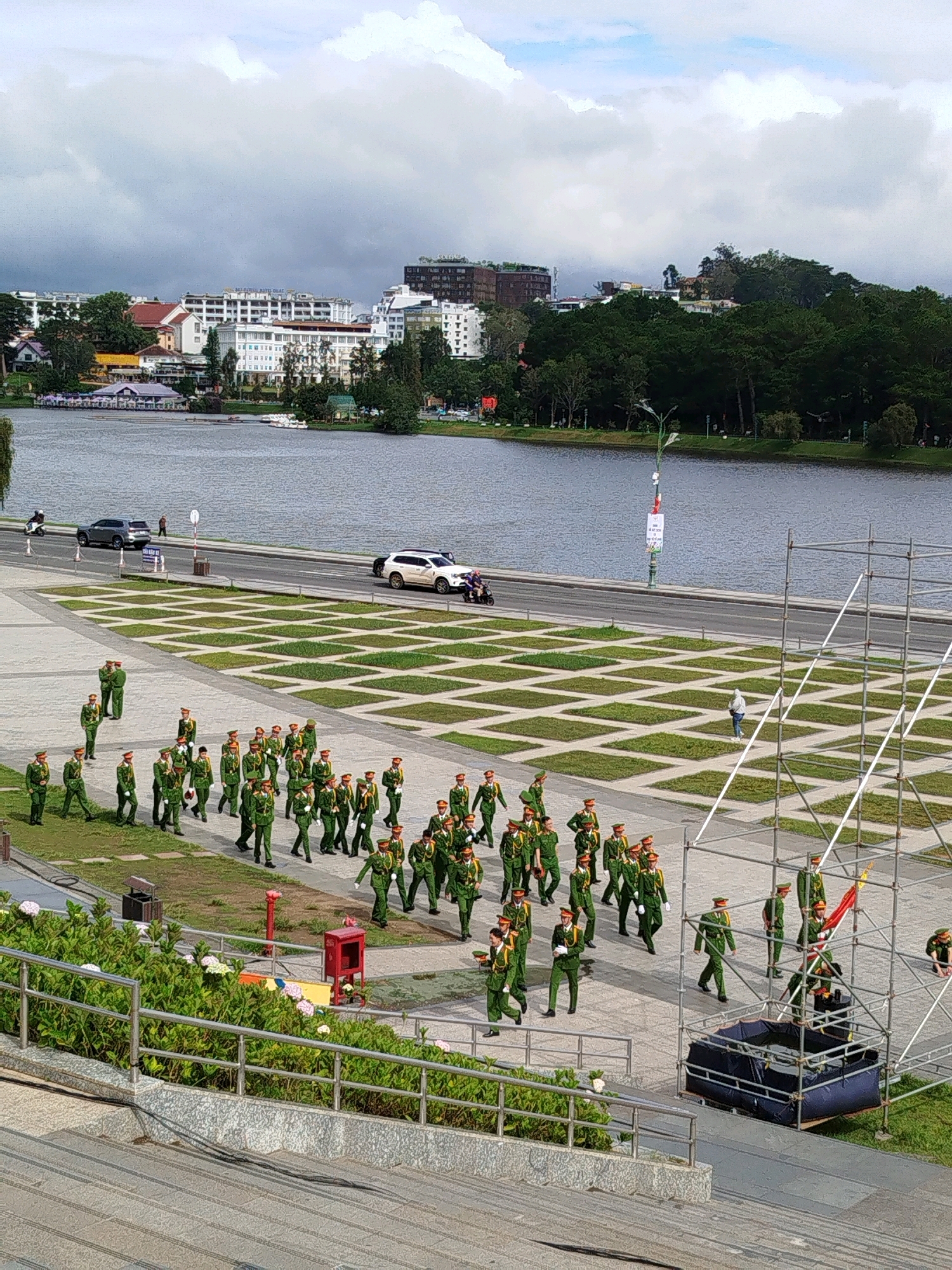 The image captures a daytime outdoor scene in Da Lat, Vietnam, featuring a parade of uniformed individuals walking in formation across a paved area with grassy squares. A large body of water, likely Xuan Huong Lake, forms the backdrop, with a cityscape of buildings and trees lining its opposite shore. Several vehicles, including cars and motorcycles, are visible on a road adjacent to the lake and parade area. Scaffolding and a large dark blue structure are present in the foreground, suggesting an event or construction. The sky is overcast with scattered clouds.