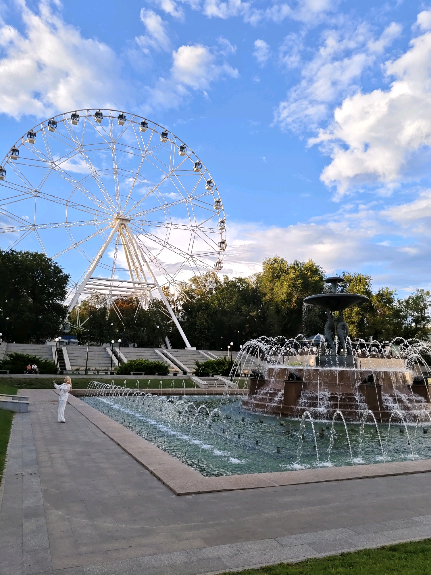 A ferris wheel and fountain are in a park in Rostov-on-Don, Russia. The ferris wheel is white with silver spokes and is partially visible against a blue sky with white clouds. In the foreground, a multi-tiered fountain sprays water into a pool, with a statue of three figures at its center. A woman in a white dress is standing on a paved pathway to the left of the fountain, taking a selfie. The park has green trees and grass, and some people are sitting on benches in the background.