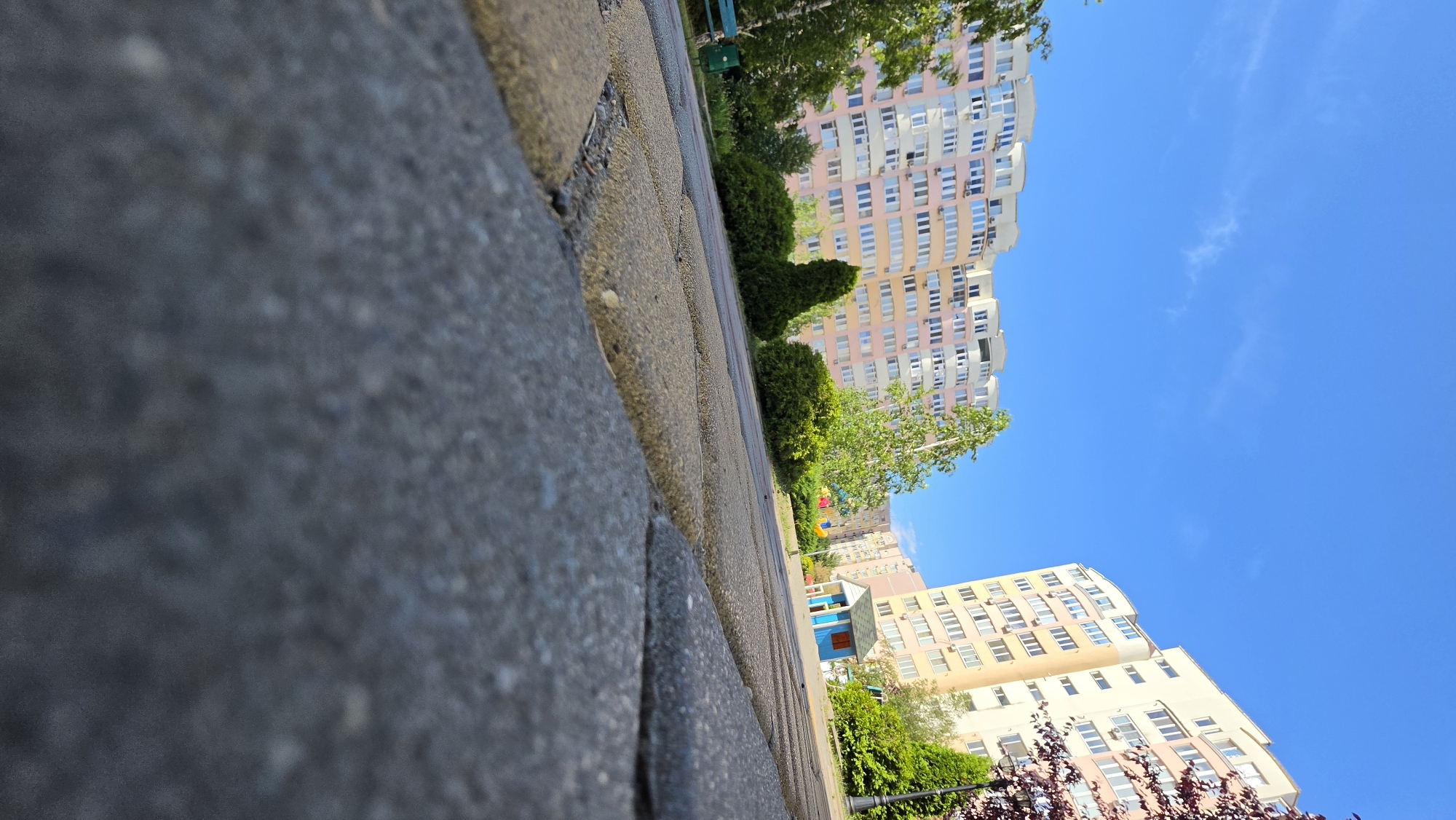 The image depicts a low-angle view of a paved walkway in front of residential buildings under a clear blue sky. The walkway is constructed from large, rectangular interlocking pavers in shades of gray and tan, with a close-up perspective on the texture of the paving stones in the foreground. Beyond the walkway, several tall, modern apartment buildings with light-colored facades and numerous windows are visible, featuring balconies and air conditioning units. Green trees and bushes are interspersed around the buildings and along the walkway, adding a touch of nature to the urban landscape. A small, brightly colored playhouse, painted blue with a red roof, is situated on a patch of grass in the middle ground, suggesting a residential area with amenities for children. The overall scene conveys a sense of a well-maintained residential complex on a sunny day.